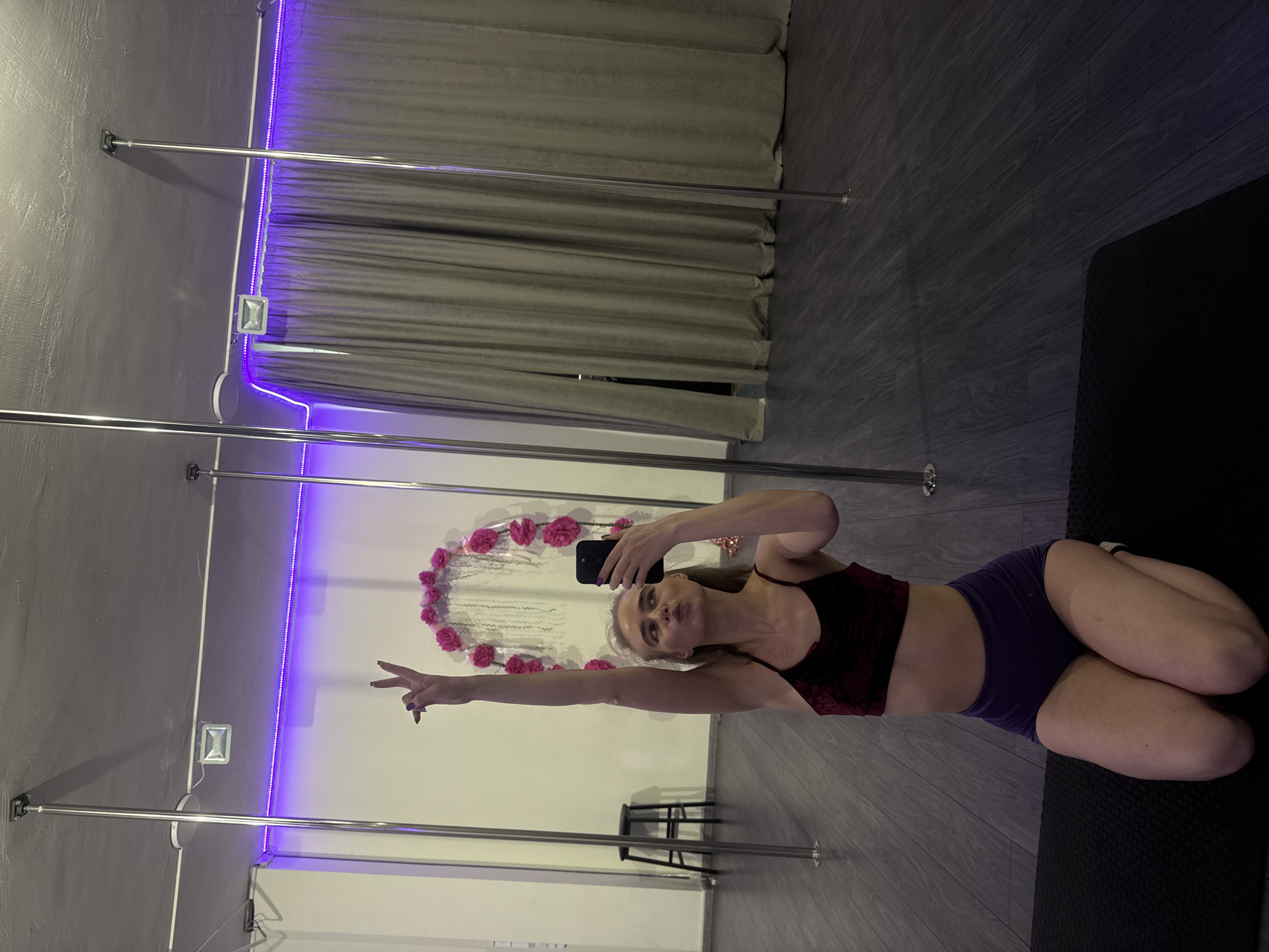 A woman is depicted in a mirror selfie, kneeling on a dark mat in what appears to be a pole dancing or fitness studio. She is facing the mirror, holding a black smartphone with her left hand to take the picture, which partially obscures her face. Her lips are pursed in a playful "duck face" expression. Her right arm is extended upwards, making a peace sign with her fingers.
She is wearing a dark, possibly velvet-textured, sports bra or crop top in a shade of maroon or dark red, paired with dark blue or purple athletic shorts. Her legs are folded beneath her as she kneels.
The studio features multiple polished silver vertical poles, characteristic of pole dancing, stretching from floor to ceiling. A large mirror covers the wall in front of the woman. Along the top edge of the mirror, a strip of vibrant purple LED lighting illuminates the space. A small, square silver floodlight is mounted on the grey wall to the left of the mirror. The floor is covered with dark grey, wood-patterned laminate or vinyl flooring.
In the reflection behind the woman, a light-colored wall is visible, adorned with a heart-shaped floral decoration made of pink flowers and white hanging elements. Further in the background of the reflection, grey curtains are drawn, and a dark stool can be seen behind one of the poles. The overall setting suggests an indoor environment, brightly lit by artificial light sources. There is no visible text in the image.