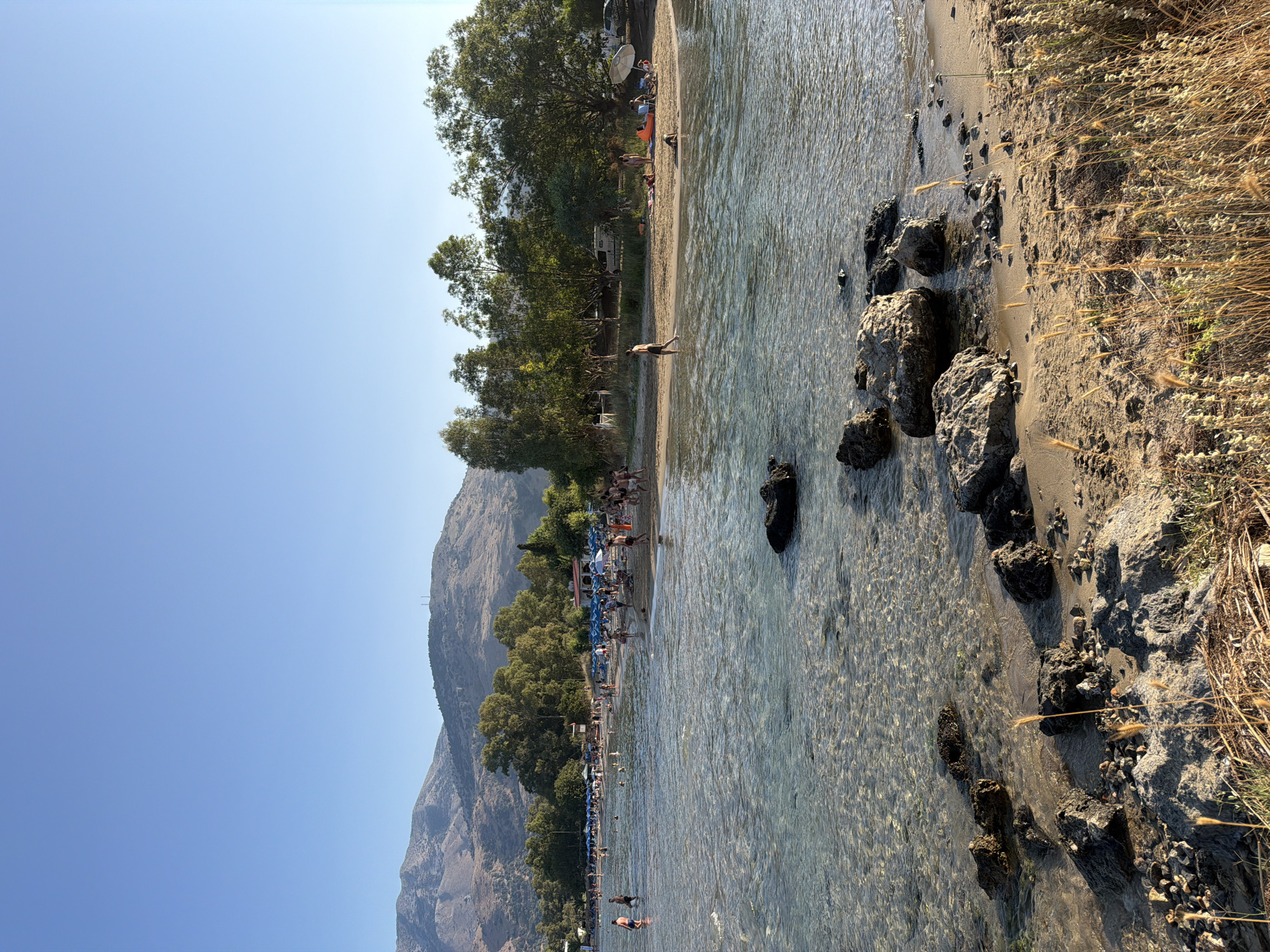 The image depicts a sunny day at a beach in Georgioupoli, Greece. The foreground features a sandy shore with scattered rocks and dry grass. The shallow, clear water of the Aegean Sea laps gently onto the beach, revealing pebbles and submerged rocks beneath the surface.
In the mid-ground, the water is populated with several individuals, some wading and others swimming. The beach is lined with numerous blue umbrellas, providing shade for beachgoers. People are visible sitting on the sand, walking along the shore, and interacting with each other. Some individuals are sunbathing or relaxing under the umbrellas.
The background is dominated by lush green trees and vegetation, with a few structures visible, possibly including small buildings or part of a resort. In the far distance, rolling hills and mountains, characteristic of the Cretan landscape, form the horizon under a clear blue sky. The overall scene conveys a relaxed and idyllic beach atmosphere.