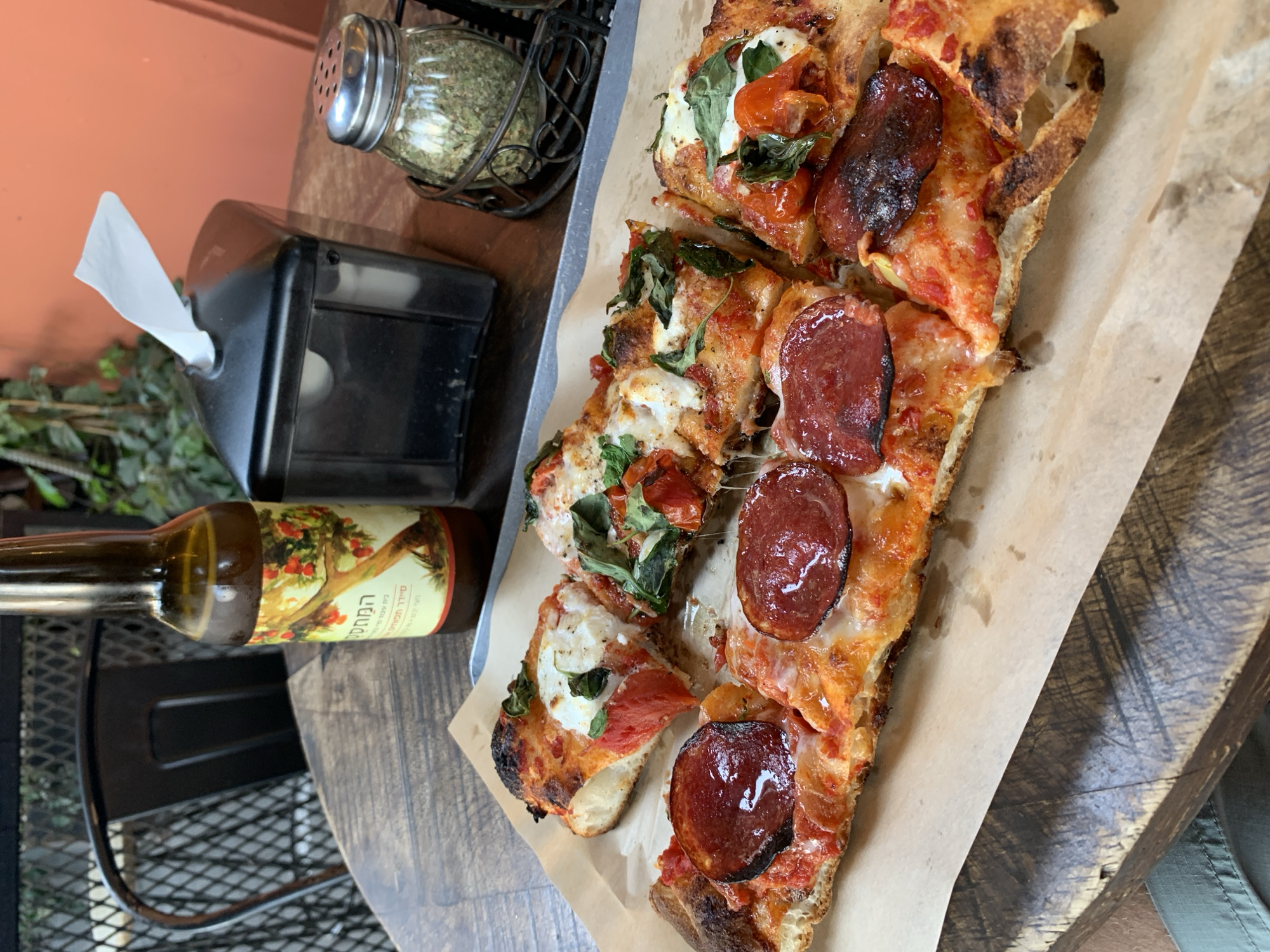A close-up, high-angle shot captures several slices of rectangular pizza served on parchment paper, resting on a metal tray on a wooden table. The pizza appears to be a thick-crust variety, possibly a focaccia or Roman style, with visible charring on the crust from a hot oven. Some slices are topped with bright red pepperoni, curled from the heat and glistening with oil, and melted white cheese. Other slices feature fresh mozzarella, halved cherry tomatoes, and vibrant green basil leaves. Streaks of melted cheese indicate its warm, freshly prepared state.
To the left of the pizza, a brown glass bottle with a colorful label depicts a tree bearing red fruit (likely olives, given the text). The label features Hebrew text, including "המחסן" (HaMachsan - The Warehouse/Store), "שמן זית כתית מעולה" (Shemen Zayit Katit Meu'leh - Extra Virgin Olive Oil), and "חומציות מרבית 0.8%" (Maximum acidity 0.8%). Behind the bottle, a black metal chair is partially visible. Further back, a black napkin dispenser with a single white napkin sticking out, and a clear glass shaker filled with dried green herbs (likely oregano), sit on the table. A green artificial plant adds a touch of greenery to the scene. The background reveals a warm, peach-colored wall, suggesting an indoor or shaded outdoor setting during daytime in Tel-Aviv, Israel. The image captures a meal being enjoyed, highlighting the appetizing pizza and accompanying condiments.