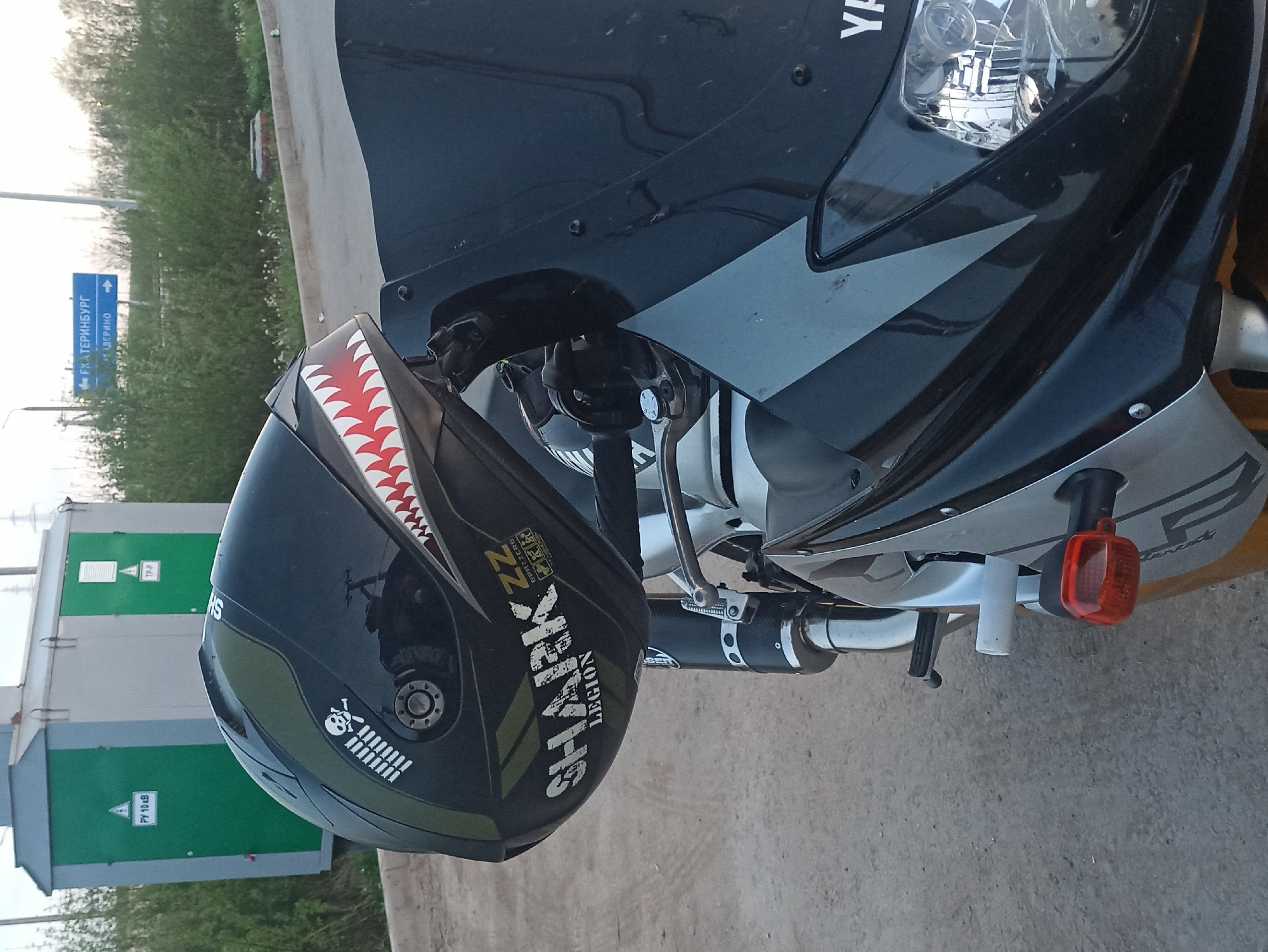 A motorcycle with a black and grey paint scheme is parked on asphalt in an outdoor setting. The front portion of the motorcycle is in focus. A black helmet with white shark teeth design and “SHARK” branding is resting on the handlebars. To the left of the motorcycle, there is a green and grey electrical substation. Behind the substation is green vegetation. To the left is a blue directional sign indicating the direction to the city of Ekaterinburg in Russian.