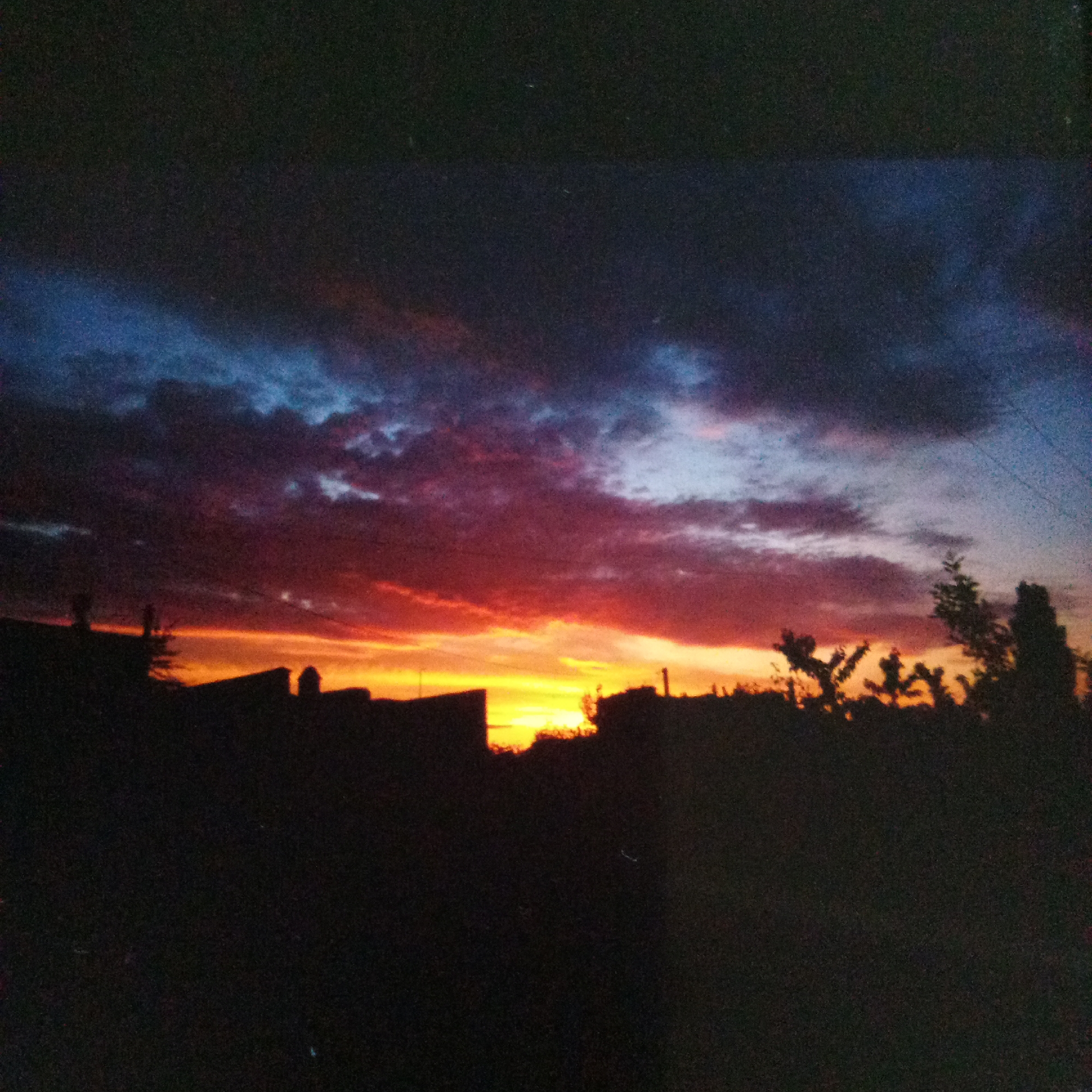 The image depicts a silhouetted landscape against a dramatic sunset sky, featuring vibrant hues of orange, red, pink, and blue. Dark clouds are present in the upper portion of the sky. In the foreground, the dark silhouettes of buildings and trees are visible, suggesting an urban or suburban setting. The overall impression is one of twilight in Myrhorod, Ukraine.
