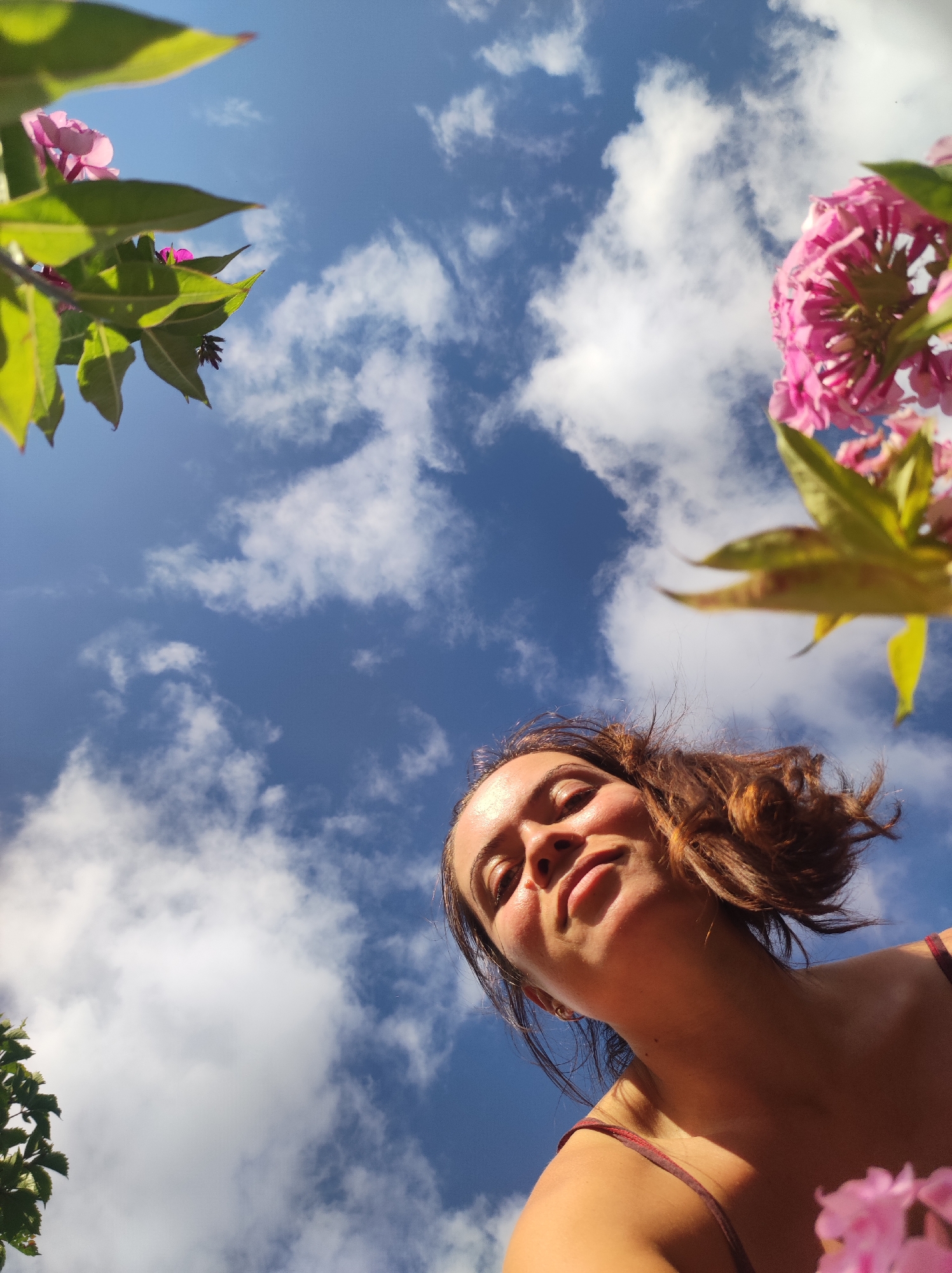 An eye-level, low-angle shot captures a young woman from the shoulders up, looking towards a bright blue sky dotted with fluffy white clouds. Her head is tilted back, and her face is illuminated by sunlight, highlighting her features and a hint of a smile. Her brown hair is slightly disheveled, framing her face. Pink flowers and green leaves are visible in the foreground, partially obscuring the sky and adding a vibrant splash of color to the scene. The overall impression is one of a person enjoying a bright, sunny day outdoors, likely in a garden or park setting. The image does not provide any specific visual cues to pinpoint its location within the city of Foky, Russia.