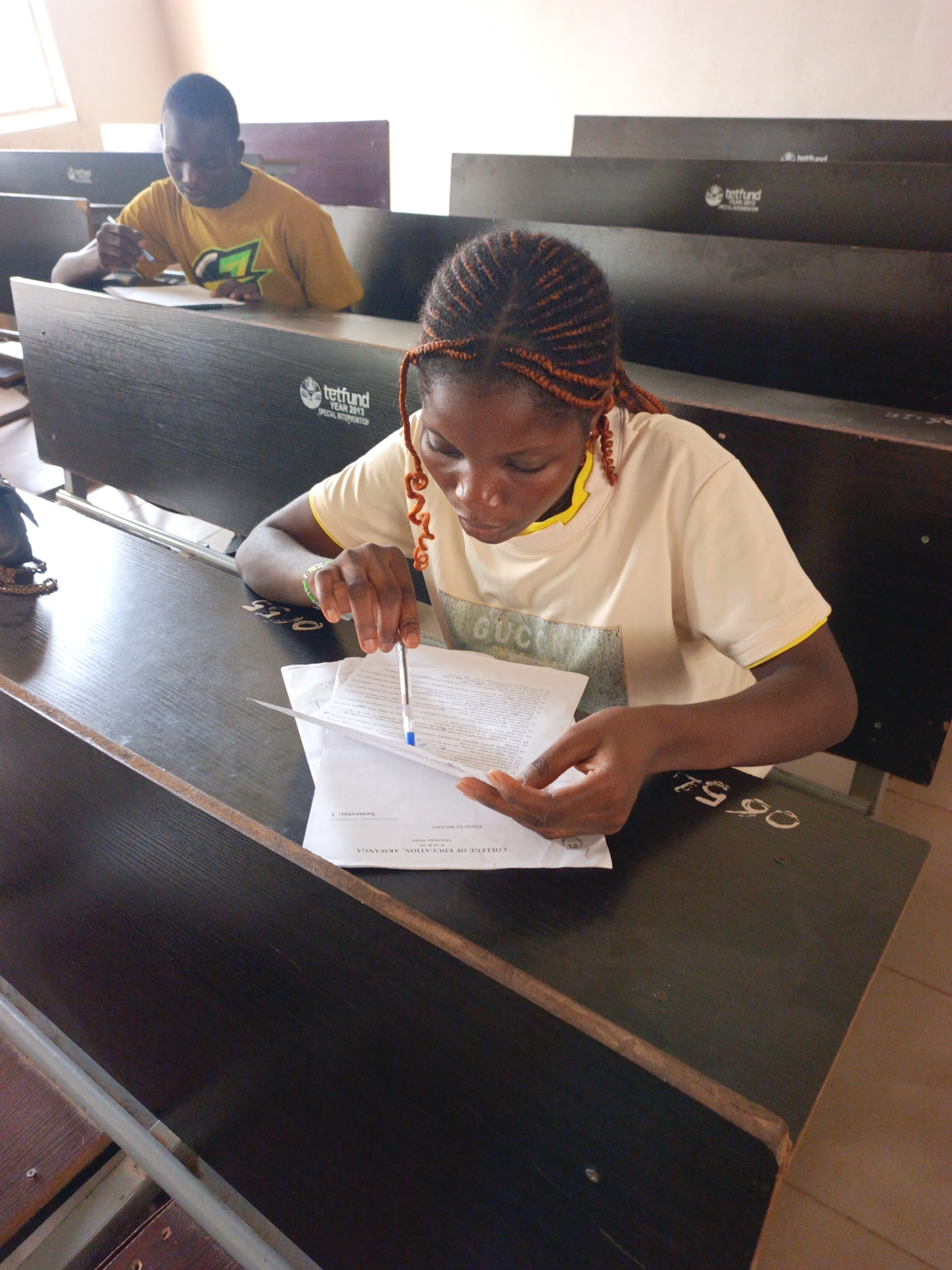 The image shows two people, presumably students, seated at desks in what appears to be a lecture hall or classroom. The scene takes place at the College of Education, Akwanga, Akwanga State, Nigeria. The first person is a young woman with braided hair seated at a desk and focused on a document in her hands, which she appears to be studying or working on with a pen. Her shirt says, “GUCCI”. The other student is a young man wearing a yellow shirt seated further back, also focused on writing or reading something. The desks are black with a metal structure, and have "tetfund YEAR 2013 SPECIAL INTERVENTION" printed on the front. The numbers "06 51" are written on the desk in the foreground. The image suggests a setting of focused study or academic activity.