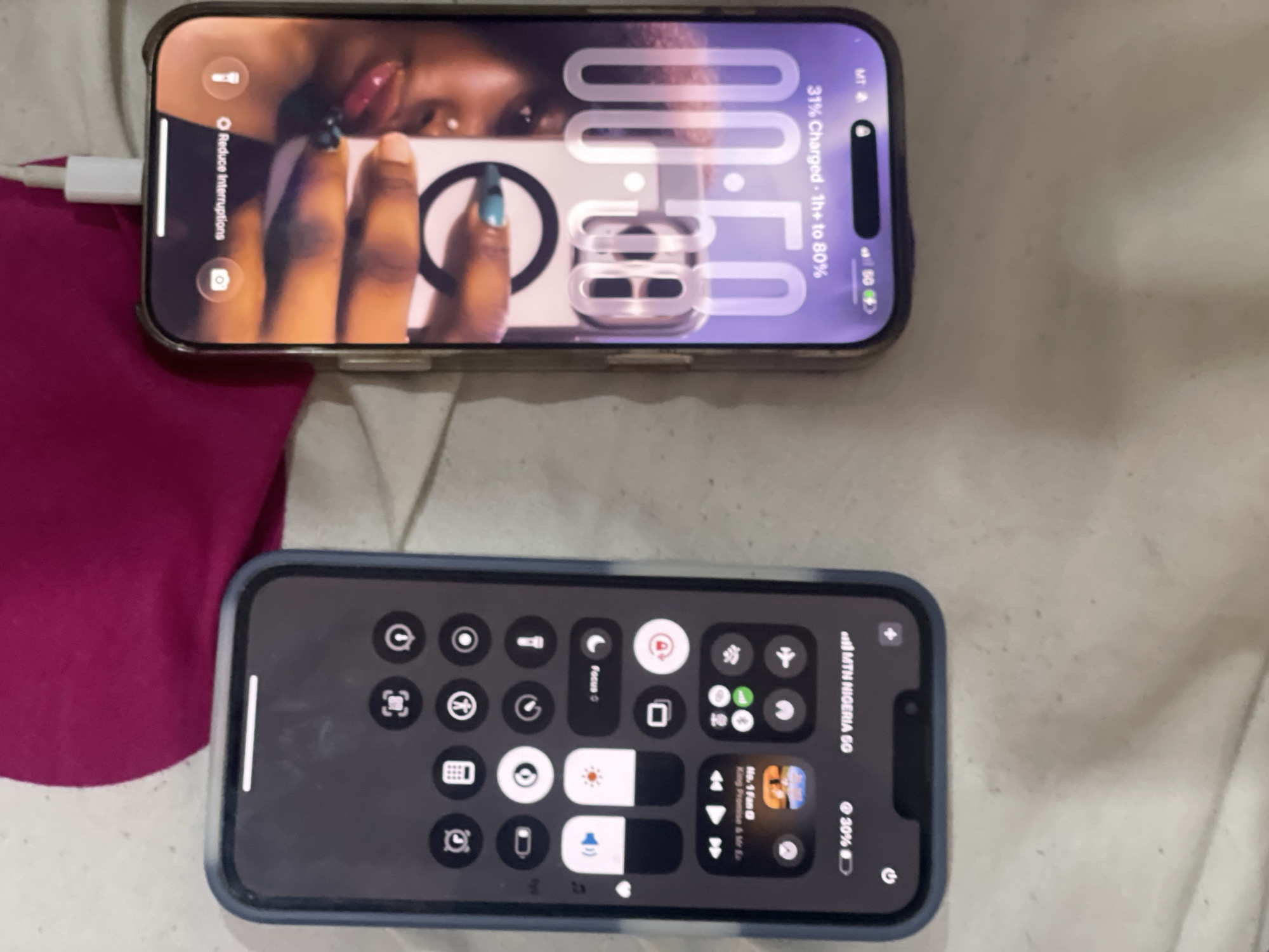 The image shows two iPhones lying on a surface, possibly a bed, with bedding visible in the background. The iPhone on top is charging and displays a lock screen with a photo of a person’s face and the time "00:50". The charging cable is connected to the phone. The iPhone below displays a control center with various settings toggles, including Focus, brightness, volume control, and a music player card with "No. 1 fan" by King Promise & Mr Eazi visible. The phone has a network provider label "MTN NIGERIA GO". Both phones are showing battery percentages of 31% and 30% respectively. The image's location is implied by the mobile network provider MTN NIGERIA, meaning the scene is in Nigeria, but it cannot be narrowed to the city of Abuja, Nigeria based solely on the information presented in the image.