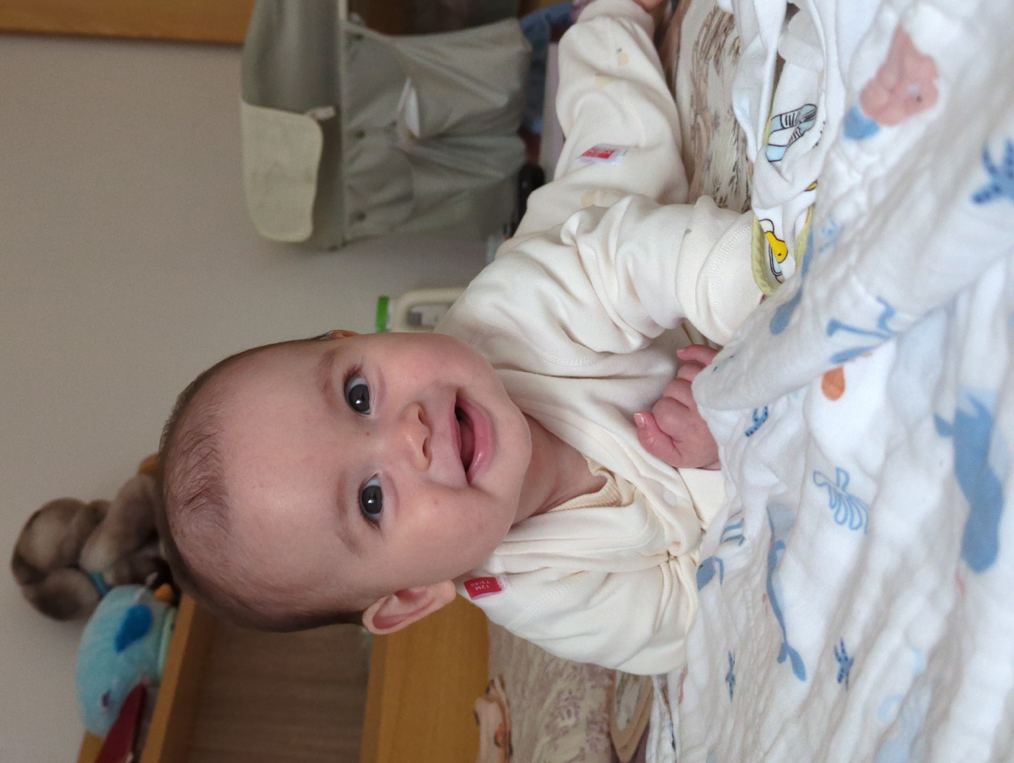 A baby lies in a crib. The baby wears a long-sleeved white outfit. The crib has a white blanket with blue designs. A stuffed animal and carrier are nearby. The location is indoors and the image's geographic location is unknown.