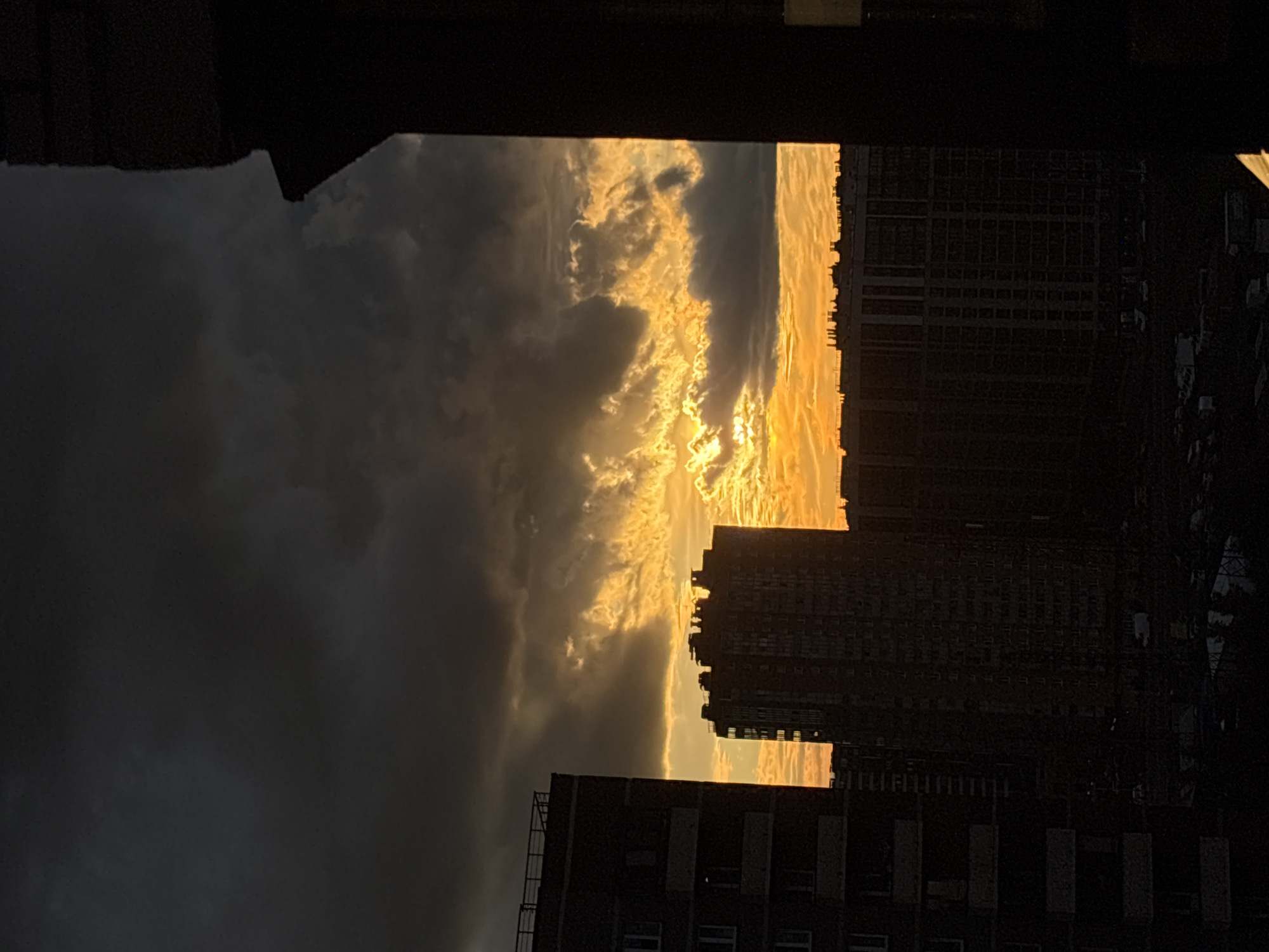 The image depicts a dramatic sunset viewed between two tall buildings. The sky is filled with dark, ominous clouds on the left, gradually giving way to brilliant golden-orange light radiating from the setting sun on the right. The sunlight catches the edges of the clouds, creating a fiery, textured effect.
Silhouetted against the vibrant sky are the dark shapes of the buildings. The one on the right is a large, multi-story structure with numerous windows, appearing as a grid pattern. The building on the left is partially visible and seems to be of similar height. The view is framed by the dark, indistinct structures in the foreground, suggesting the photograph was taken from within another building or from a rooftop.
The time of day is clearly sunset, indicated by the intense coloration of the sky and the low angle of the light. The weather appears to be mixed, with both dark storm clouds and bright, sunlit areas. The overall mood is one of awe and perhaps a touch of foreboding due to the contrast between the dark clouds and the brilliant light, creating a sense of natural drama. There is no visible text in the image. The scene is urban, likely within a densely populated area, given the presence of the tall buildings.