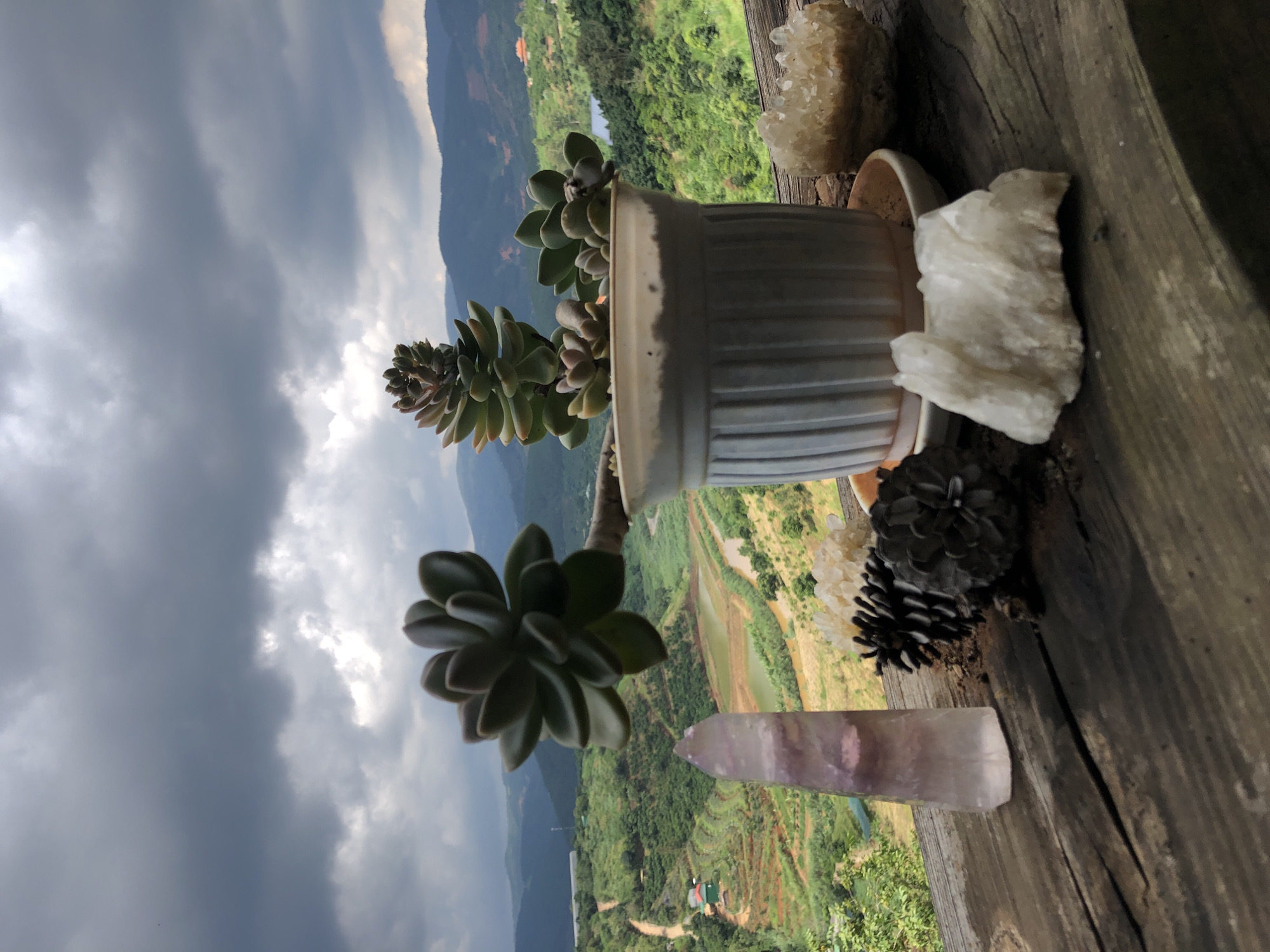 In Thôn 3, Vietnam, a weathered wooden surface displays an assortment of objects against a dramatic backdrop of cloudy skies and rolling green hills. A white ceramic pot contains two succulent plants, their fleshy leaves reaching upwards. To the left of the pot, a tall, translucent purple crystal stands upright, its facets catching the light. Adjacent to the crystal are two dark, textured pinecones. To the right of the pot, a cluster of small, clear crystals rests on the wooden surface. In the background, a vibrant landscape unfolds with terraced fields, a winding road, a body of water, and distant mountains, all under a heavy, overcast sky.