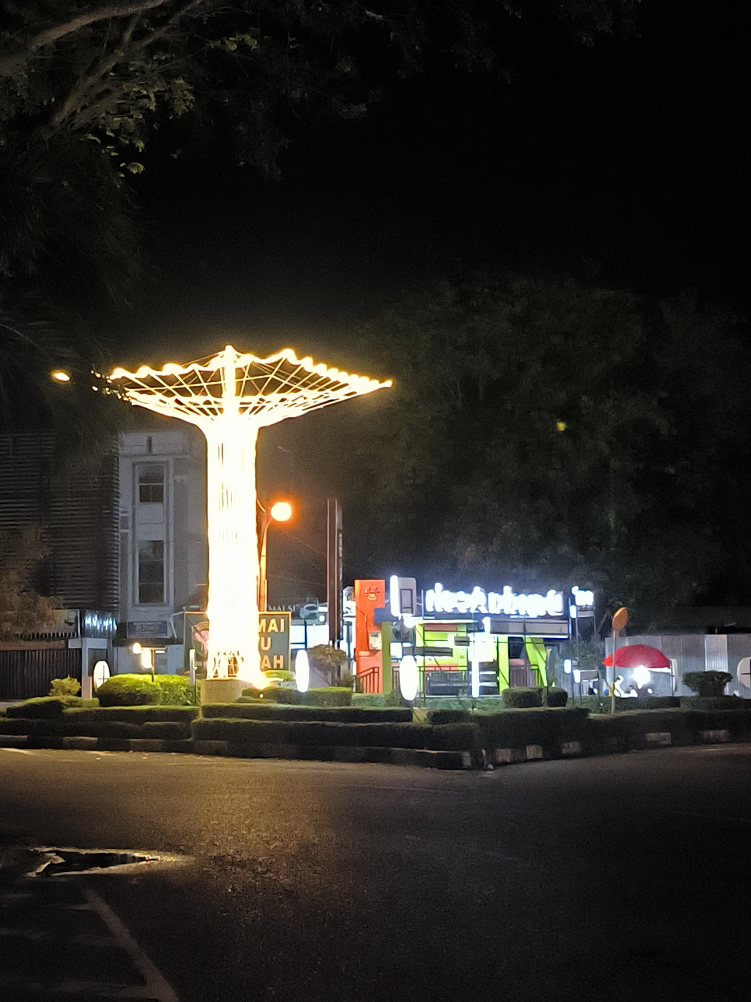 This nighttime image captures a vibrant urban scene, likely a public square or roundabout. The central focus is a large, stylized monument resembling a tree or an umbrella, brightly illuminated with numerous golden-yellow lights. This structure stands on a central island surrounded by manicured low hedges and a paved road, where a reflective puddle is visible in the foreground.
In the background to the left, a building with multiple windows can be seen. To the right, several commercial establishments are present, marked by brightly lit signs. One sign features stylized, inverted text that is difficult to decipher, possibly "e.h.d.aeki". Further right, a red umbrella is set up, beneath which indistinct figures can be faintly seen, suggesting people are gathered. Dark foliage from trees frames the top and right side of the image against the deep night sky.
The prominent monument features text at its base, which appears to read "MAI JUAH" (or "AH" for the second word). The scene is illuminated entirely by artificial lights, creating a lively and welcoming ambiance characteristic of an urban area after dark. The location is consistent with a public space in Banda Aceh, Indonesia.