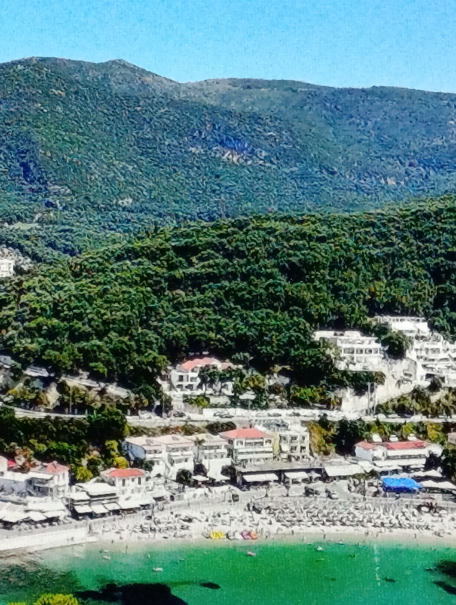 This image depicts a coastal scene. A mountain covered in green vegetation forms the backdrop. In the mid-ground, white buildings with red roofs cascade down the slope towards a beach. The beach is populated with people, some lying on the sand and others in the water. The water near the shore is clear green. The sky is blue and cloudless.