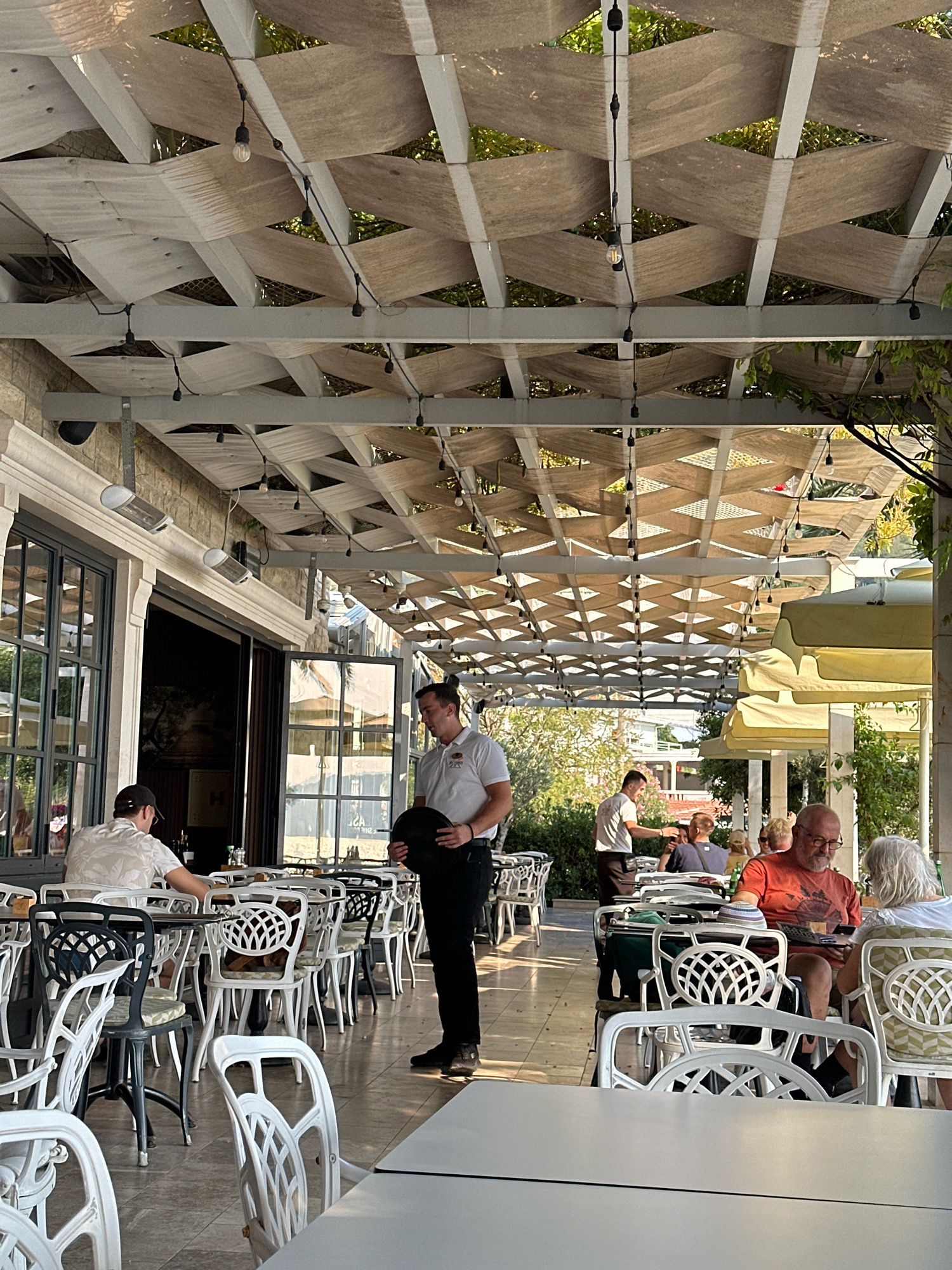 The image shows an outdoor seating area of a restaurant in Bečići, Montenegro, covered by a white lattice structure with beige fabric panels and string lights. Several tables and white ornate chairs are arranged on a tiled floor. A waiter in a white shirt and black pants, holding a black hat, stands in the foreground. To the left, a man wearing a patterned short-sleeved shirt and a baseball cap sits at a table. In the background, other patrons are seated at tables, and some are being served by another waiter. The scene suggests a pleasant daytime setting, with greenery visible in the background.