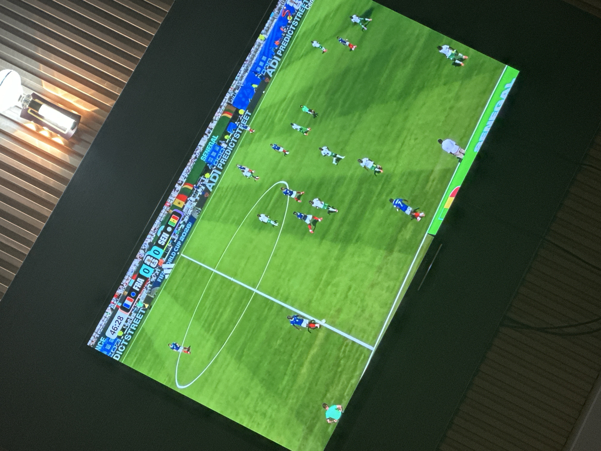 The image shows a television screen displaying a soccer match. The score is 0-0. The time is 46:28. Players in blue and white jerseys are on the field. There are advertisements visible on the screen, including "ADI PREDICTSTREET". The soccer field is green with white lines. In the background, there are spectators in the stands. The television is placed on a dark surface, possibly a table, with vertical wooden panels visible above and behind it. The lighting in the room appears to be dim, with a bright light fixture visible in the upper right portion of the image. The image quality is slightly blurred due to the angle and possible movement. The scene is indoors, and there is no specific information in the image that places it in Rayfield, Nigeria.