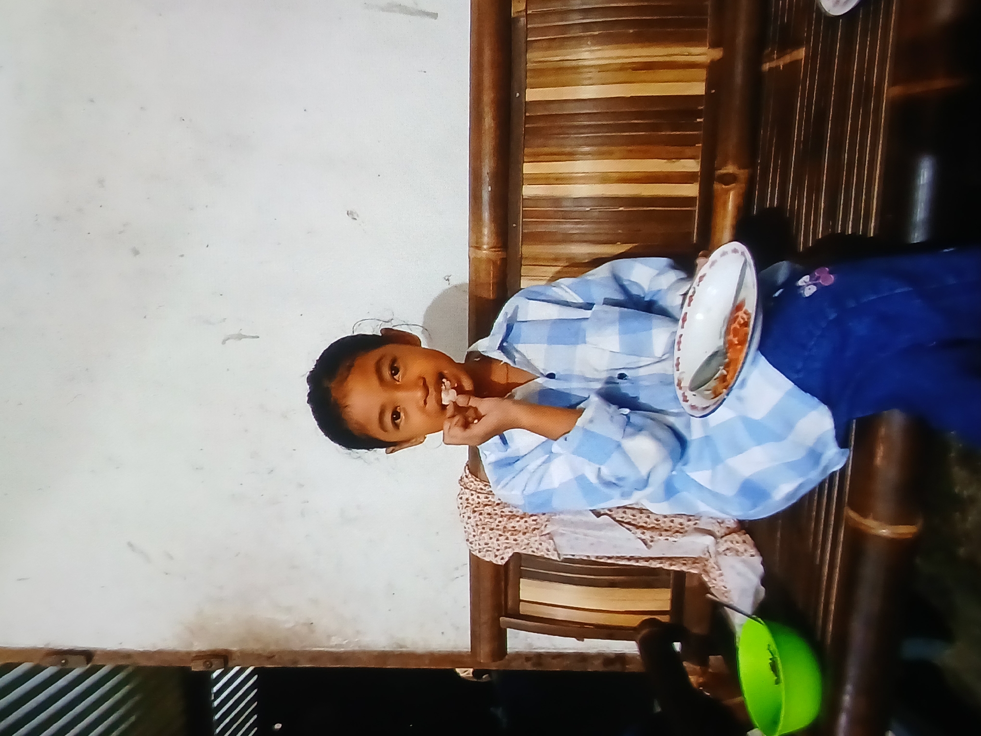 A young girl is seated on a bamboo bench, eating. She is wearing a blue and white checkered shirt and dark blue pants with a flower applique on the right leg. In her right hand, she holds a piece of food to her mouth. She is holding a white plate with a red rim in her left hand. The plate contains food and a fork. To the left of the girl is a green bowl filled with green leafy vegetables. The bench is situated against a white wall, and to the left of the girl, draped over the back of the bench, is a patterned cloth. The overall environment suggests an outdoor or semi-outdoor setting, possibly in the city of Setia Bakti, Indonesia.