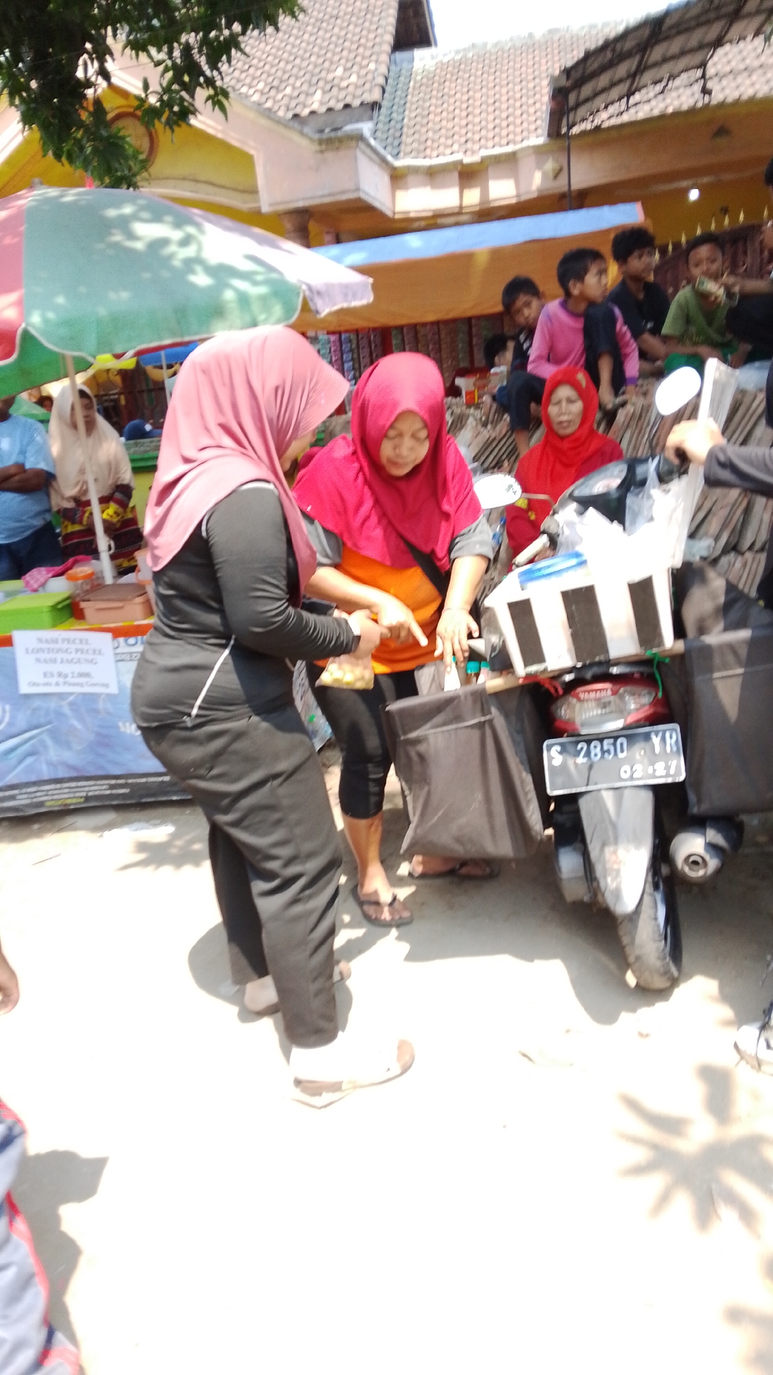 In Mancar, Indonesia, a scene unfolds outdoors, possibly at a market or roadside stall. Two women wearing hijabs are the central figures. One, in a pink hijab and black outfit, stands with her back to the camera, holding a bag of what appears to be food items. The other woman, in a bright red hijab and an orange and grey top, is bent over, pointing and interacting with the first woman. A parked motorcycle with a white box on the back and black saddlebags stands to their right. The motorcycle's license plate reads "S 2850 YR 02-27". In the background, other people, including children, are visible, some standing near a stall with colorful awnings. The ground is paved, and shadows suggest it is a sunny day. A sign in the foreground is partially visible with Indonesian text including "NASI PECEL" and prices.