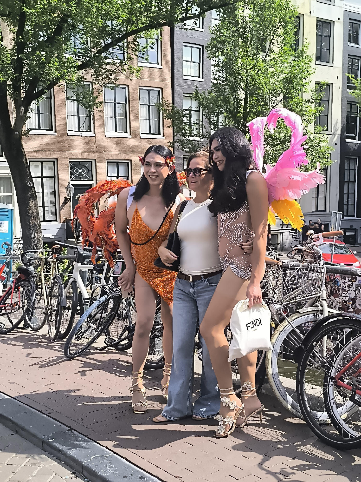 Three people are posing for a photo in front of a canal in Amsterdam, Netherlands. The person on the left is wearing an orange sequined dress and large orange feather accessories. The person in the middle is wearing a white tank top and blue jeans. The person on the right is wearing a light-colored, sparkling leotard with large pink and yellow feather wings. They are all smiling and looking at the camera. In the background, there are bicycles parked along the canal and traditional Dutch buildings. The street is paved with brick, and shadows are cast by the buildings and trees. A red car is visible in the distance. The overall scene suggests a festive or celebratory event, likely a parade or festival given the costumes.