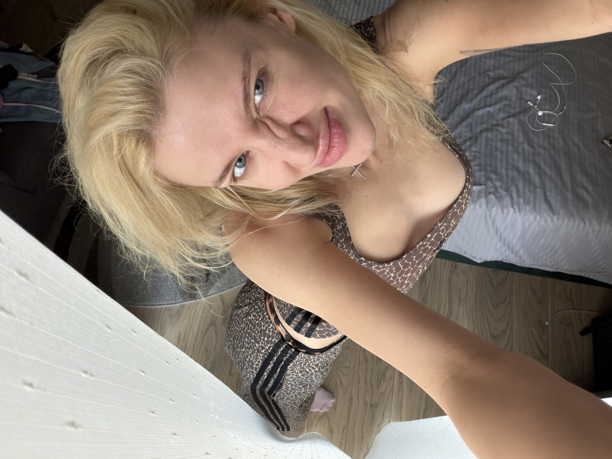A blonde woman with blue eyes takes a selfie indoors. She wears a spotted print top and matching pants with three black stripes down the sides. A silver necklace is visible at her neck. Behind her is a grey bed with headphones and a black object. A white wall or door is to her left, and a hardwood floor is beneath her. The location in Belgrade, Serbia, cannot be determined from the image.