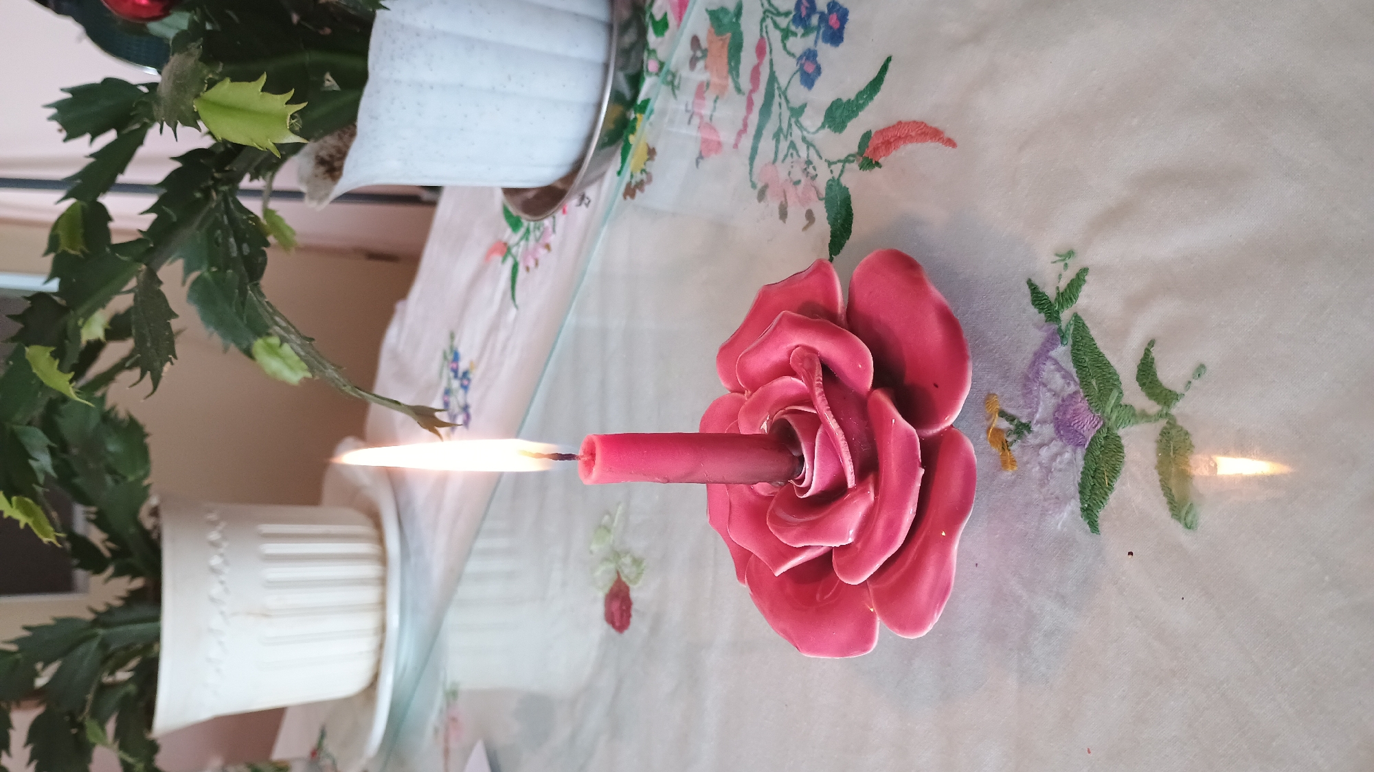 The image depicts a still life composition on a glass table covered with a white embroidered tablecloth. In the foreground, a pink candle stands upright in a pink ceramic rose-shaped candle holder. The candle is lit, and its flame is visible. The tablecloth features floral embroidery in various colors, including green, purple, and blue. In the background, two potted plants with green leaves are placed on white pots, one on the left and another on the right. The overall scene suggests an indoor setting, likely a home environment. The location in Sokolov, Czechia, is not discernible from the visual elements in the image.
