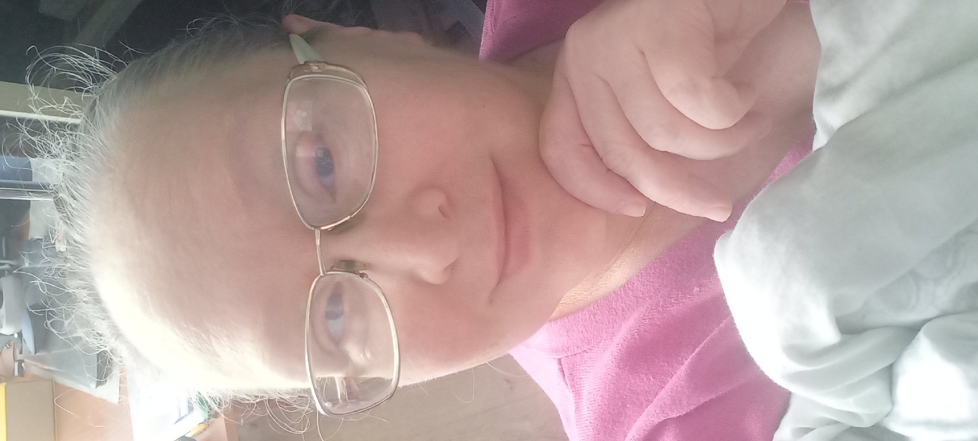 A close-up, slightly angled, portrait-style photograph shows a person with fair skin and light blonde or gray hair. The person is wearing glasses with gold-colored frames. The person's hand is positioned near their face. The person is wearing a pink garment, and a white fabric is visible to the right. The photograph is taken indoors. It is not possible to determine the location of the image as Izhevsk, Russia.