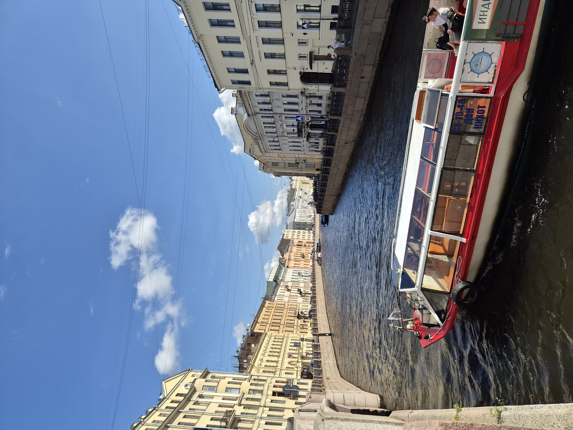 In a city canal, a red and white tour boat with a glass roof navigates the dark water under a blue sky with scattered clouds. Several people are seated on the upper deck of the boat, enjoying the view. The canal is bordered by stone embankments and lined with colorful, multi-story buildings in various shades of yellow, orange, and white, showcasing architectural details like arched windows and decorative facades. The scene appears to be a sunny day in a European city, likely a tourist destination.