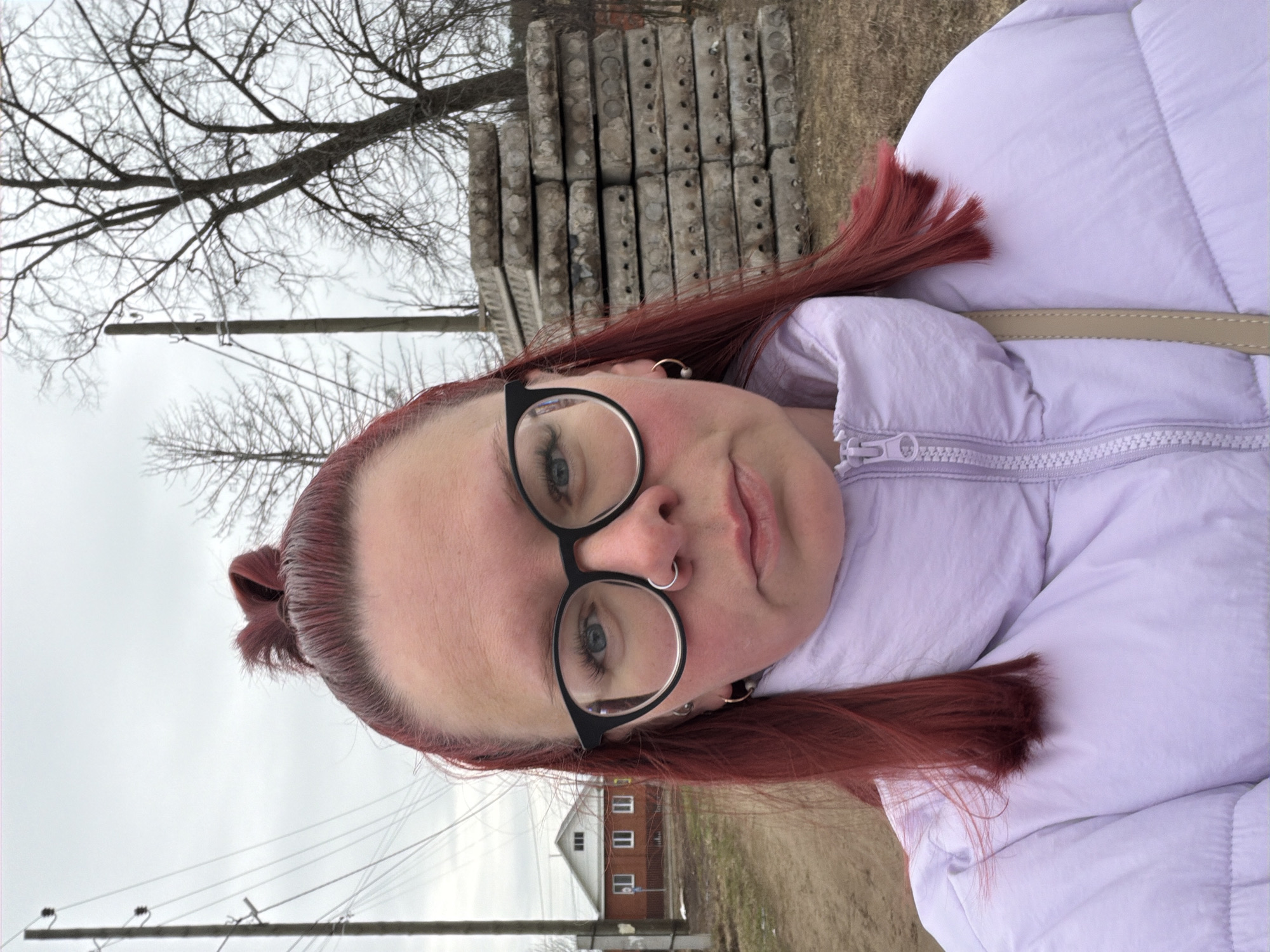 A young woman with red hair, wearing round glasses and a light purple puffer jacket, is taking a selfie outdoors in Chaikovsky, Russia. She has a nose ring and is wearing earrings. The background includes a stack of concrete slabs, a leafless tree, utility poles with wires, a house with white windows, and a dirt path. The sky is overcast.