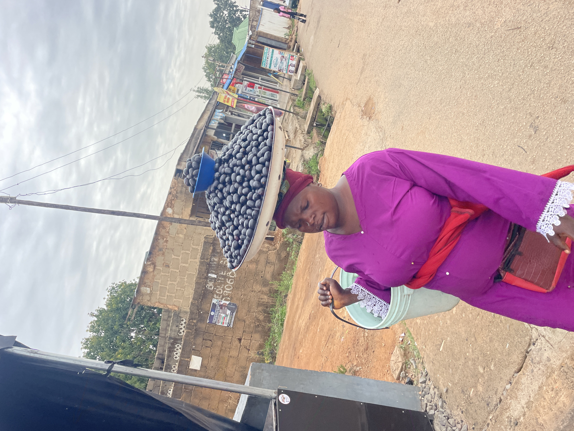 An image shows an elderly woman carrying a large tray of dark-colored fruits on her head, presumed to be agbalumo or African star apples. The woman is dressed in a purple outfit with white lace trim. A red sash is draped across her shoulder, and she carries a light-colored bucket in her hand. A building is visible in the background with various shop signs, including one for modifications and painting. The background suggests an urban setting, consistent with the street scenes in the city of Jos, Nigeria. The image is somewhat tilted, which makes it appear as if everything has been set on an angle.