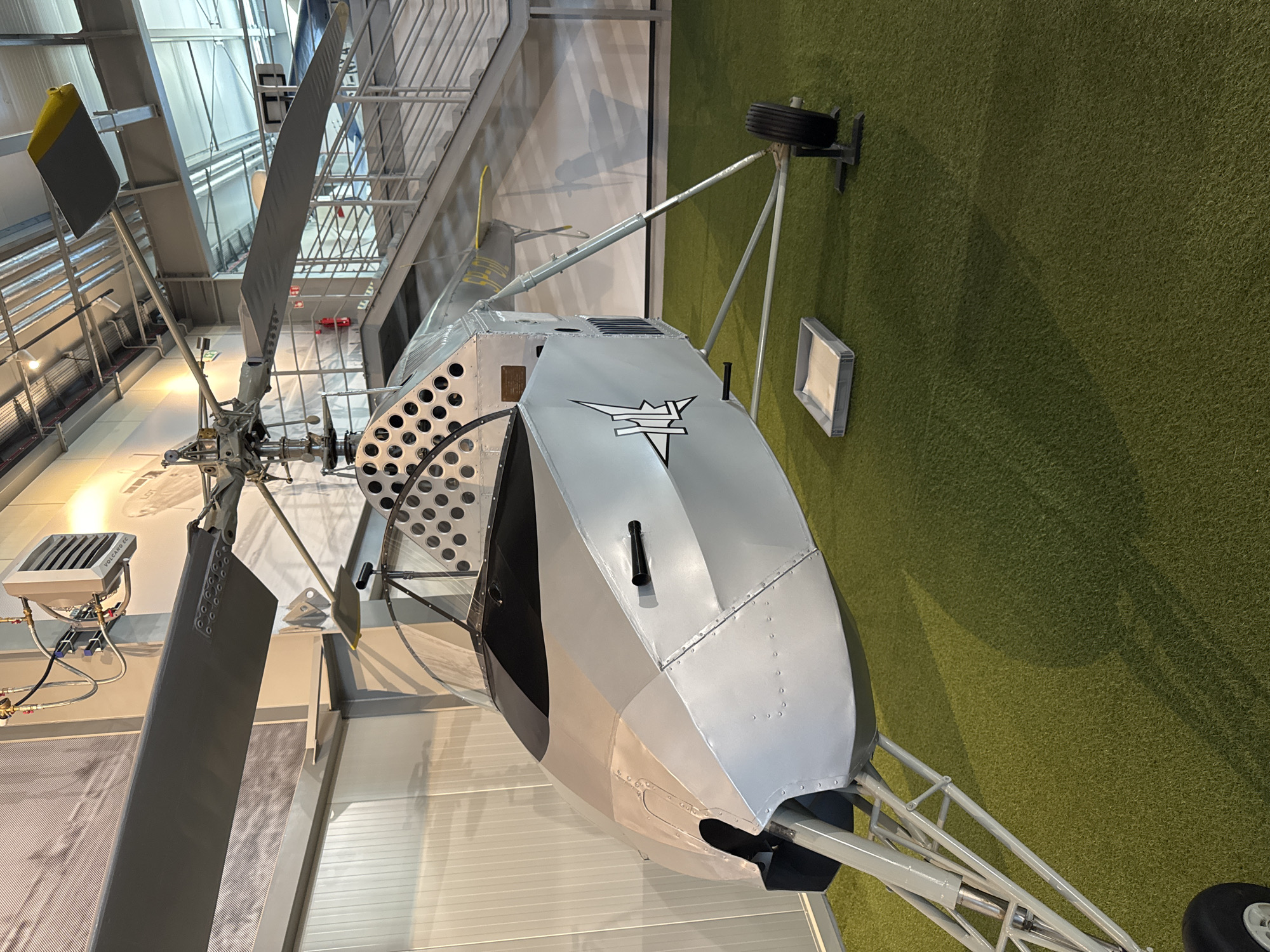 The image shows a close-up, eye-level view of a light gray helicopter on display, situated on a patch of artificial turf that resembles grass. The helicopter is positioned at an angle, with its nose pointing towards the left side of the frame. The main rotor blades are visible, extending upwards and to the left. The cockpit features a black canopy. There are various metal components and structural elements visible, including landing gear and a tail boom. A logo, a stylized white star with wings, is prominently displayed on the side of the helicopter's fuselage. In the background, there is a gray staircase with metal railings and a light-colored wall with vertical paneling. A white ventilation unit is mounted on the wall above the helicopter's rotor. The scene appears to be indoors, possibly in a museum or exhibition hall, given the lighting and display environment.