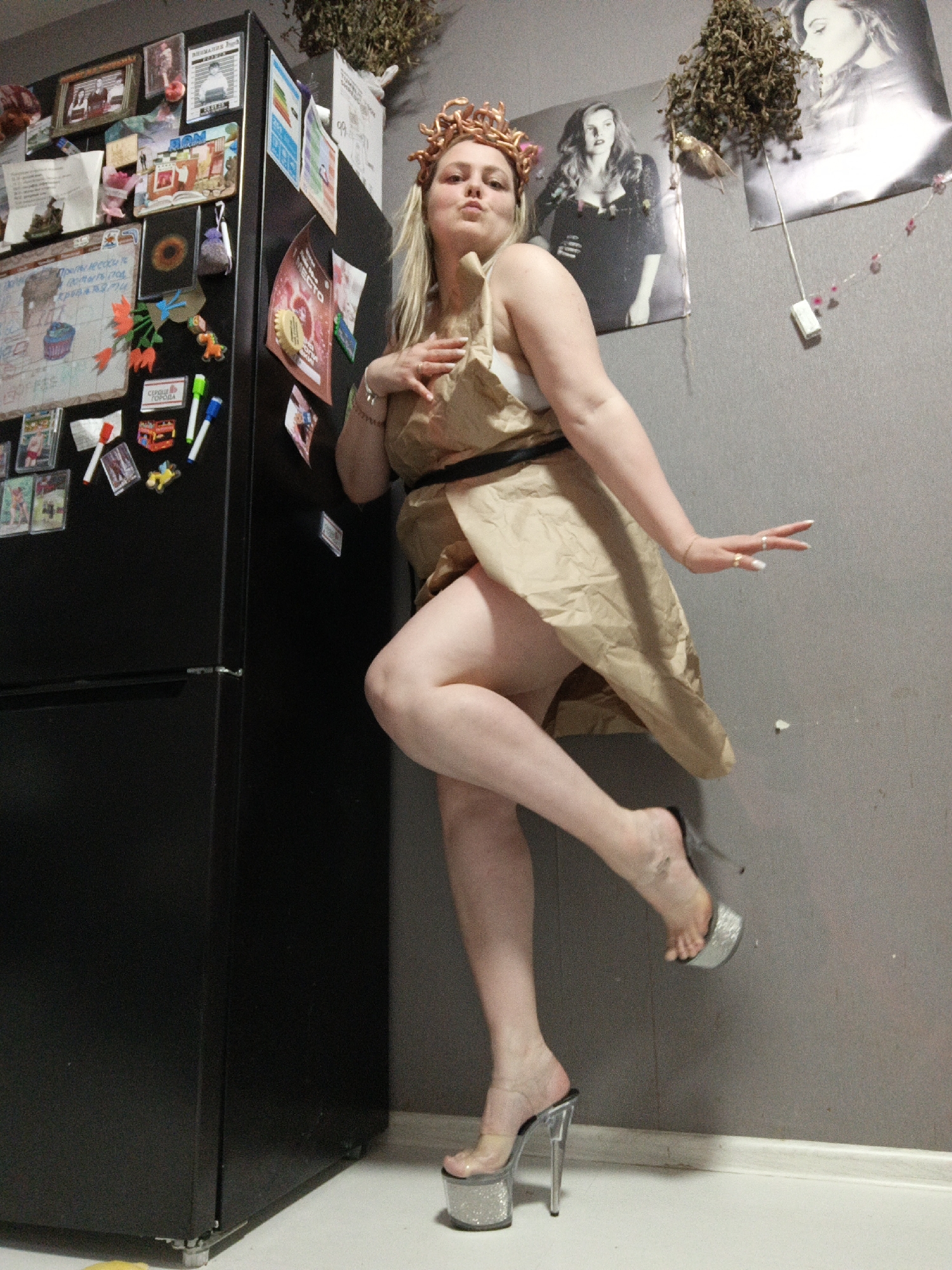 A young woman is captured in a dramatic pose within an indoor setting, likely a home in Markovskiy, Russia. She stands with one leg lifted, leaning against a black two-door refrigerator that is heavily adorned with various magnets, papers, and small decorative items.
The woman has blonde hair and is wearing a distinctive light-brown, paper-like dress belted at the waist, revealing a white bra underneath. A crown-like headpiece with snake-like elements, possibly Medusa-inspired, adorns her head. She wears very high, clear platform heels with glittery soles. Her fingers display rings, and her fingernails are long and painted. Her expression is serious with pursed lips, contributing to the theatrical nature of her pose.
The background features a plain gray wall decorated with several posters, including two large black and white images of women, one of whom resembles Adele. Dried floral or herbal arrangements hang from the wall, and a white electrical outlet is visible. The floor is light-colored with a simple baseboard.
Visible text on the refrigerator includes a heart-shaped magnet with "СЕРДЦЕ ГОРОДА" (Heart of the City) in Cyrillic, and a poster with "МЕСТО" (Place) in Cyrillic. Other indiscernible Cyrillic writings appear on various notes and papers affixed to the fridge. The scene suggests a creative or costumed photoshoot taking place in a personalized domestic environment.