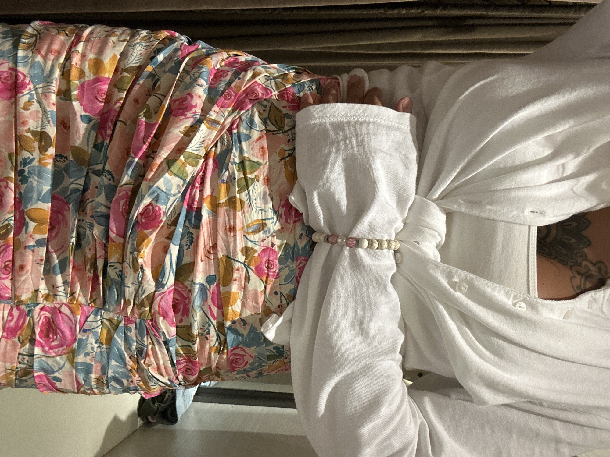 A person, whose arm is visible, is wearing a floral dress and a white shirt. The dress has a pleated skirt with a colorful floral pattern featuring pink roses, blue flowers, and yellow accents. The white shirt is buttoned up and has a sleeve tied with a beaded bracelet. The background appears to be indoors, with a textured surface resembling blinds or a slatted wooden structure. The lighting suggests it is daytime. The overall impression is a close-up shot focusing on the clothing and the arm of the person. There is no visible text in the image.