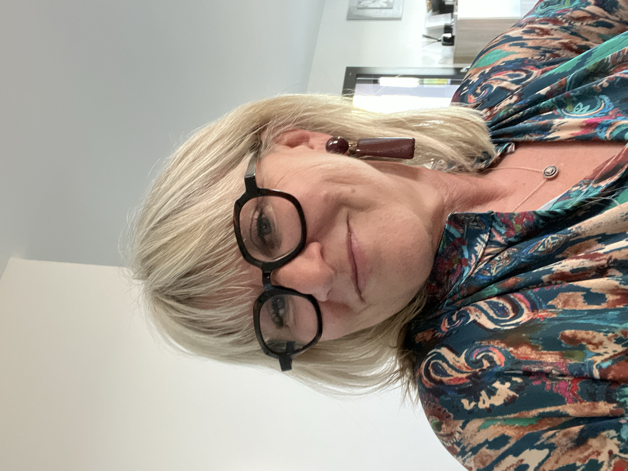 A middle-aged woman with blonde hair and glasses is looking at the camera. She is wearing a colorful blouse and a necklace. She is indoors. The location is inferred to be in or around Chambœuf, France based on the provided information.