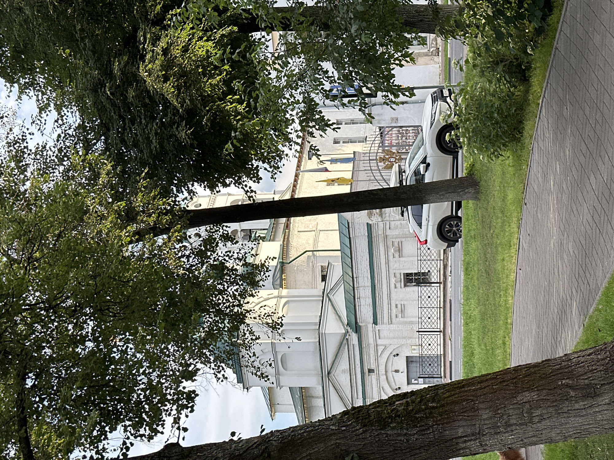 A white SUV with a roof rack is parked on the side of a road next to a paved walkway and a grassy area. In the background, a white building with green trim and a green dome is visible. Several trees with green foliage are in the foreground, obscuring parts of the building. The sky is partly cloudy. The location is Putyatino (Semivragovskaya S/A), Russia.