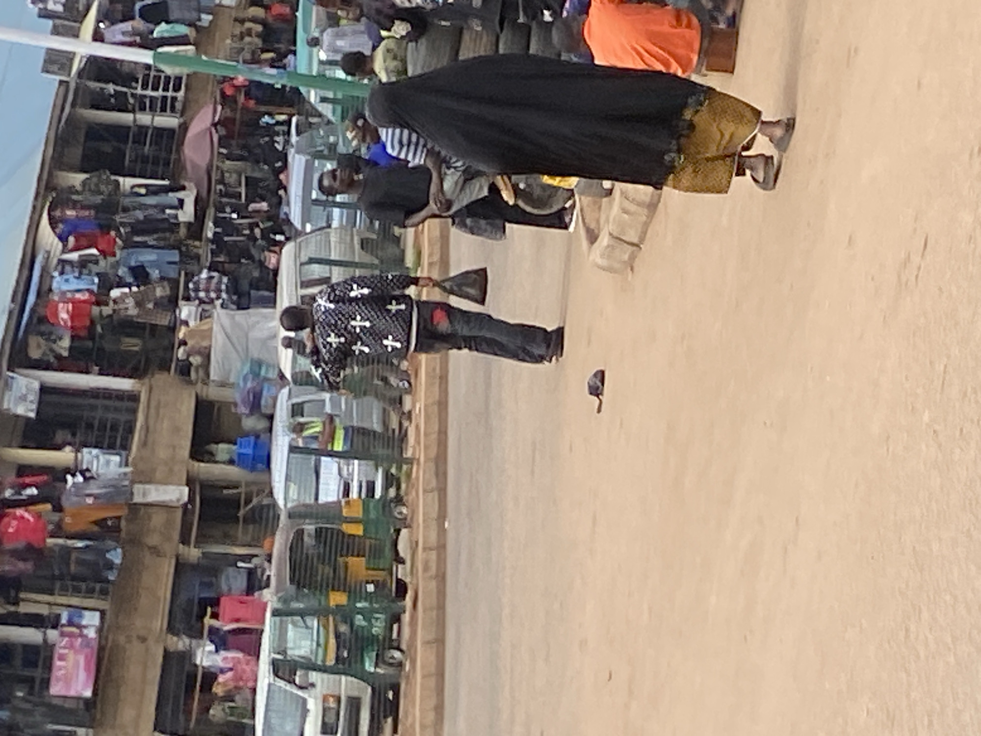 This image captures a bustling street scene in Bukuru, Nigeria, likely a market area, during daylight hours. The picture is rotated 90 degrees counter-clockwise, showing a dirt or unpaved road with market stalls lining the left side.
In the foreground, a man wearing a black long-sleeved shirt with white cross patterns and dark pants walks towards the right side of the frame, holding a dark object or bag in his left hand. Further into the middle ground, a woman dressed in a black abaya or hijab and a golden-patterned skirt walks in the same direction, wearing sandals. To her left, another man in a black shirt and sunglasses is visible, walking in the opposite direction. A small, dark-colored pigeon is seen on the ground between these individuals.
The left side of the scene is dominated by numerous market stalls, multi-story structures made of wood and metal, filled with various merchandise. Many folded clothes of different colors (red, blue, green, white, black) are visible on display in these stalls, suggesting they are clothing shops. Several vehicles, including green and yellow tricycle-like vehicles (locally known as Keke Napep or tuk-tuks) and some white vans, are parked or moving in the background, further indicating a commercial area.
The ground is light-colored and appears dusty or unpaved, consistent with a busy market street. The bright lighting suggests it is mid-day, with no discernible shadows indicating early morning or late afternoon. There is no clearly visible text on signs or vehicles. The overall scene depicts everyday life and commerce in an African market setting.