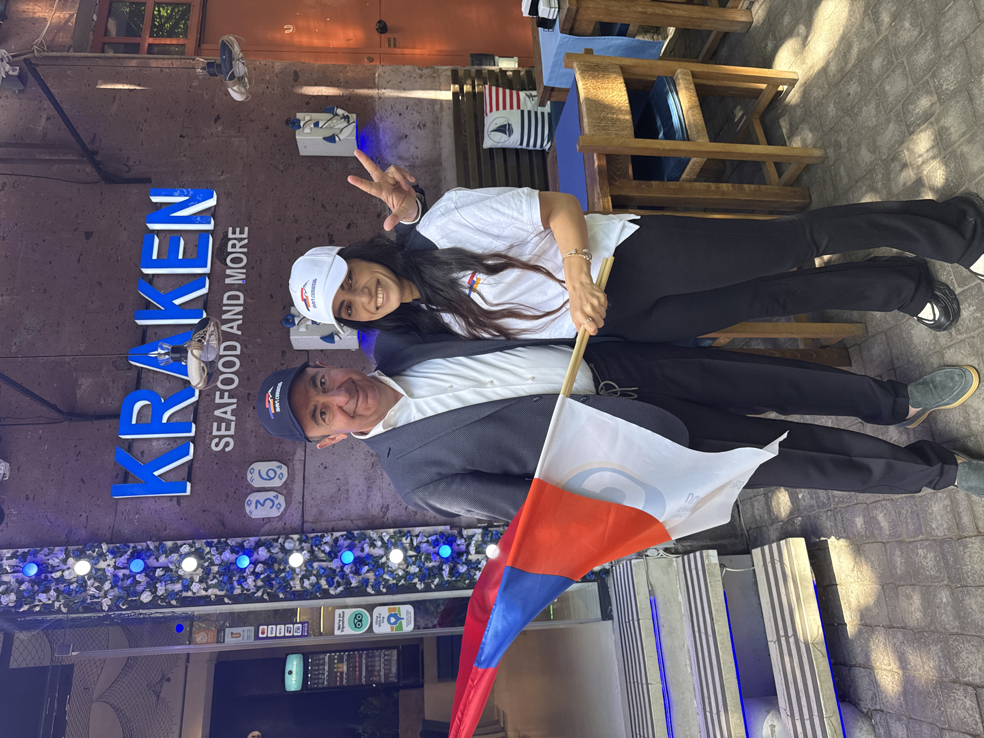 Two individuals, a man and a woman, stand outside a restaurant in Yerevan, Armenia. The restaurant has a sign that reads "KRAKEN SEAFOOD AND MORE." The man is wearing a dark suit jacket, a white shirt, and a baseball cap. He is holding a flag with white, red, and blue stripes and an emblem. The woman wears a white t-shirt, black pants, and a white baseball cap with a logo. She is holding a small stick, possibly for the flag. She is smiling and making a peace sign with her fingers. The restaurant's facade is visible in the background, with decorative lights. The scene appears to be a casual, celebratory moment captured in front of the establishment.