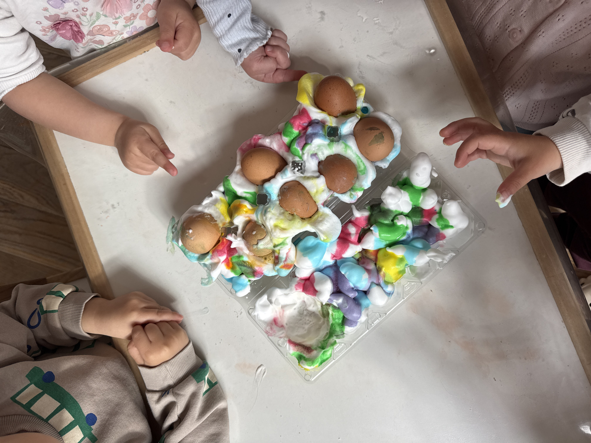 The image shows a group of children gathered around a white table or surface, interacting with an egg carton filled with eggs and a colorful mixture resembling shaving cream or foam, possibly dyed with food coloring. Four children's hands are visible, suggesting there are at least four children present. The hands are interacting with the contents of the carton, with some appearing to be poking or touching the eggs and colored foam. The eggs are brown. The overall scene suggests a child-friendly activity or art project. The image does not contain information to confirm it was taken in Sofia, Bulgaria.