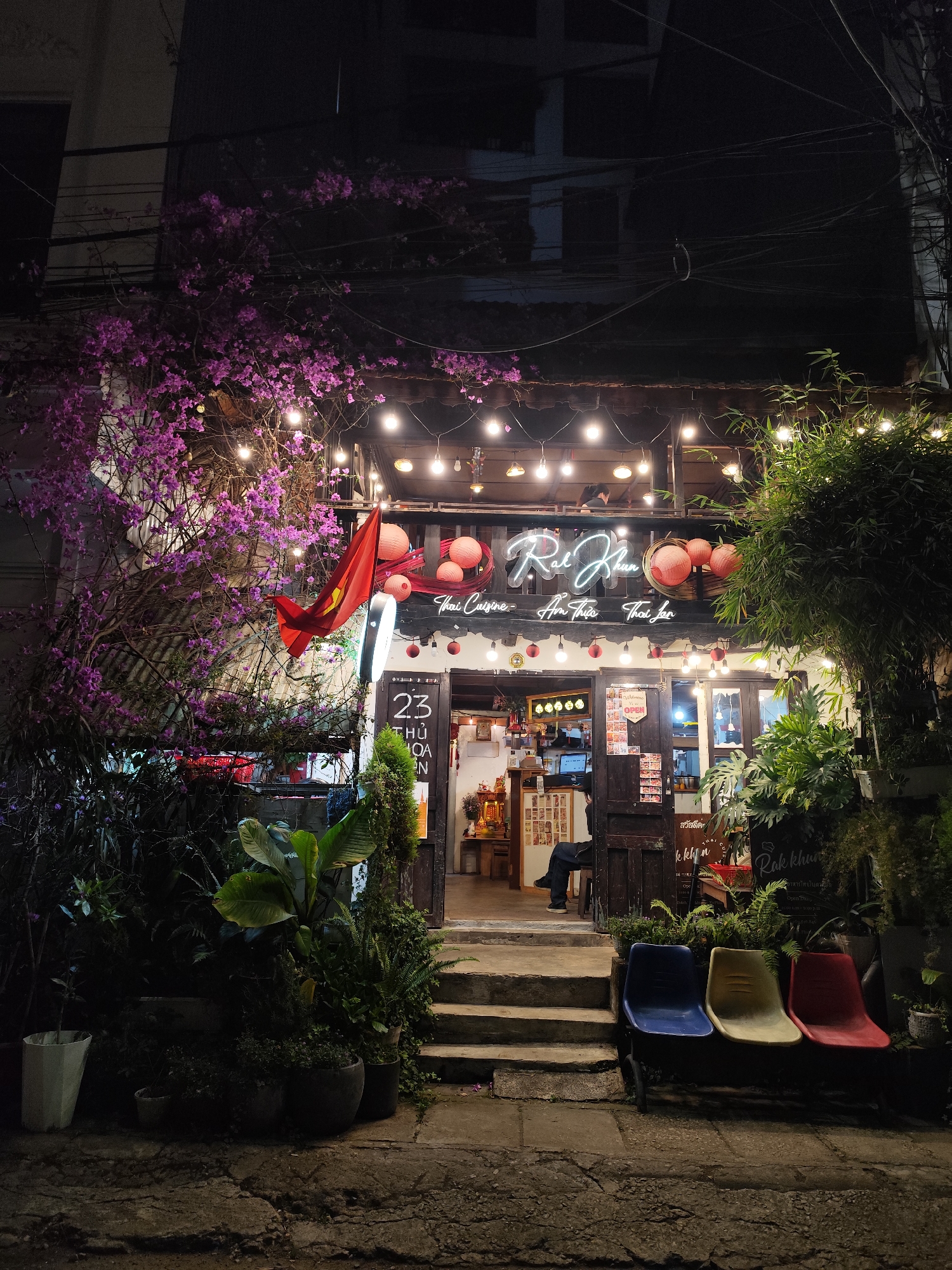 The image depicts the exterior of a restaurant called "Rak Khun" located at 23 Thu Hoa in Da Lat, Vietnam. The entrance, framed by lush foliage including flowering bougainvillea on the left and various potted plants, is illuminated by string lights and lantern decorations. A Vietnamese flag is displayed prominently to the left of the entrance. The restaurant sign, "Rak Khun," is written in neon lettering above the door. Inside, a person is visible sitting. Outside, three plastic chairs are arranged near the base of the steps leading up to the restaurant. Signage and text in Vietnamese and Thai, along with the word "OPEN," are visible on the entrance door. The setting is at night, with the restaurant’s lights contrasting against the darkness.