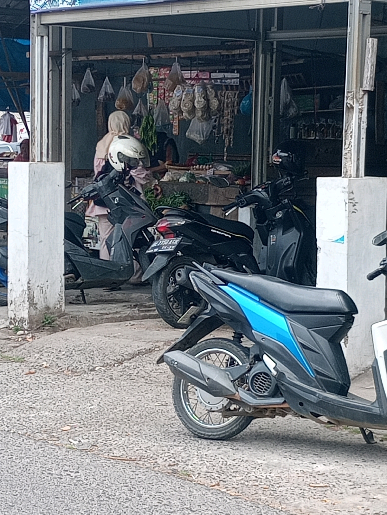 The image depicts a street-level view of a small market stall in Bandar Lampung, Indonesia. The stall is open-fronted, with shelves displaying various goods, including what appear to be packaged food items and produce. Two individuals are visible within the stall, one wearing a light-colored headscarf and a helmet, and the other partially obscured. In front of the stall, several motorcycles are parked. The foreground features a gravelly road surface. The overall scene suggests a typical day at a local marketplace in Indonesia.