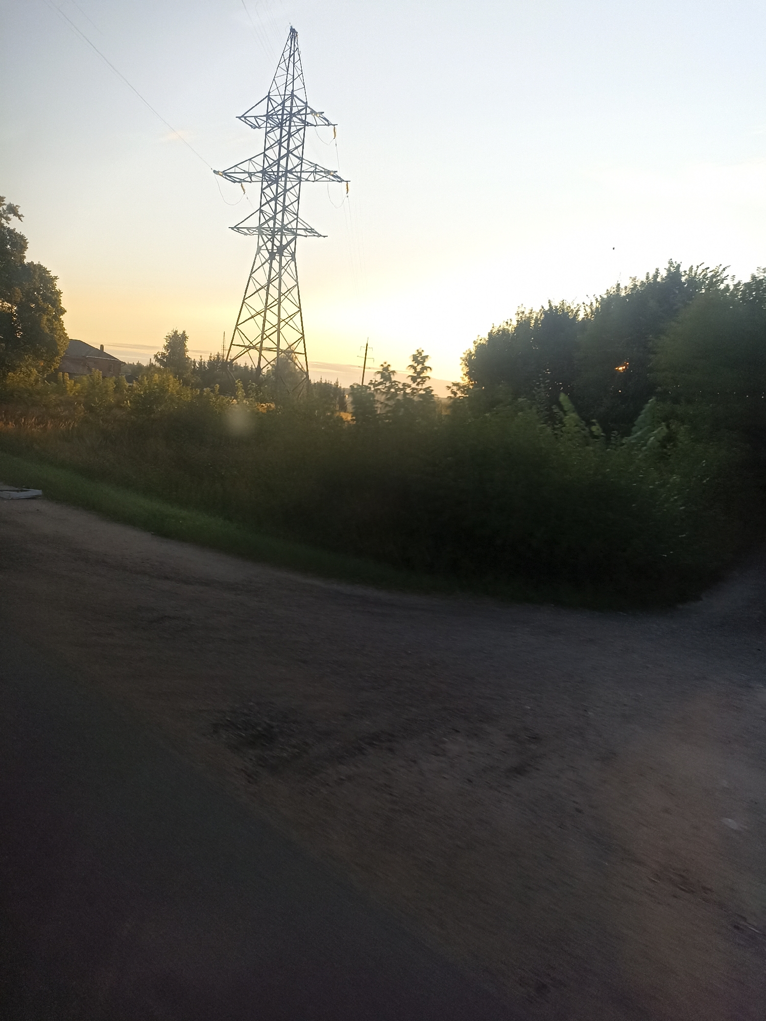 A wide-angle shot shows a power line pylon in silhouette against a pale yellow sky, indicating it is either dawn or dusk. The pylon is tall and latticed, with power lines extending from it and disappearing out of frame. To the left of the pylon, a residential building is visible, partially obscured by trees. The foreground is dominated by a dirt road, which appears to be a path or unpaved road, with some sparse vegetation along its edges. The background is a dense line of trees and foliage. The scene appears to be set in a rural or semi-rural area, possibly on the outskirts of Chekhov, Russia, given the presence of a residential building and natural landscape.