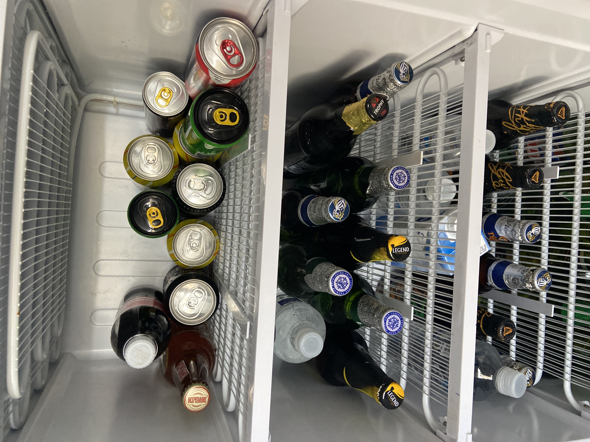 The image shows the inside of a refrigerator with multiple shelves filled with various beverages. The top shelf contains several cans of drinks, some black, some green, and some yellow. There are also two glass bottles, one dark brown and one dark red, with white and red caps respectively. The middle shelf is occupied by several bottles of water and what appear to be beer bottles with distinctive labels. The bottom shelf also holds a collection of beer bottles, some with gold caps and others with silver caps adorned with blue and white logos. The refrigerator appears to be located in a home or a commercial establishment within Gura Top, Nigeria. There are no people visible in the image.