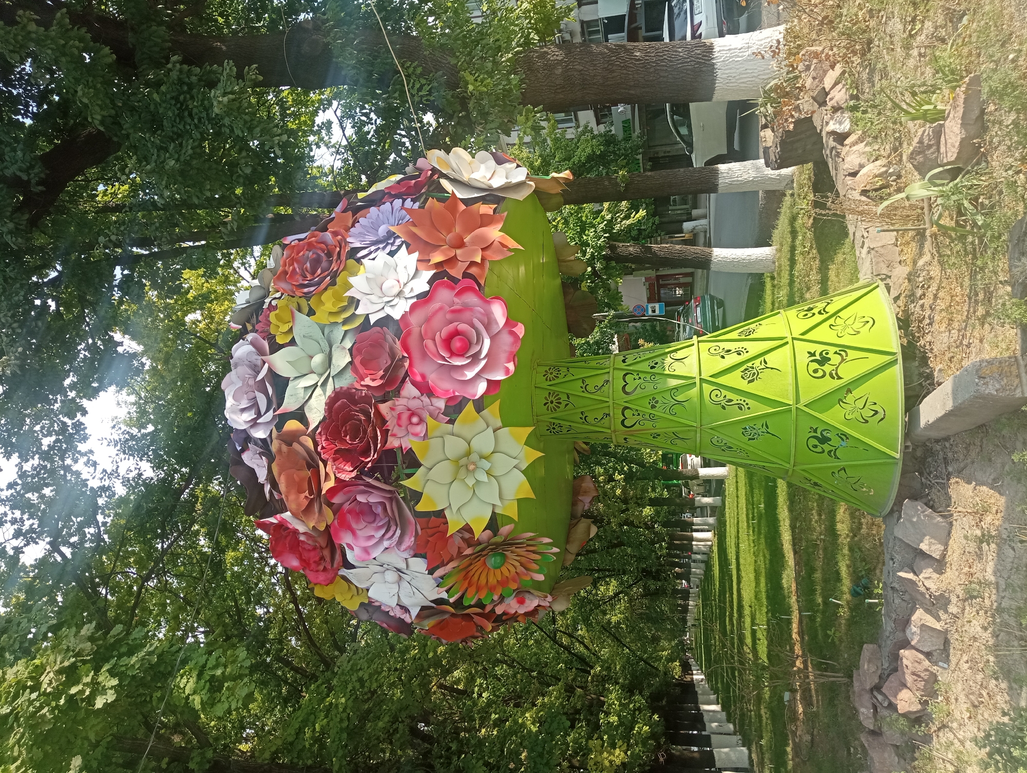 The image shows a large, decorative sculpture of a bouquet of oversized paper flowers, displayed on a light green, patterned base. The sculpture is located outdoors, in what appears to be a public park or boulevard in the city of Shymkent, Kazakhstan. The flowers are colorful and varied in size and type, with prominent red, pink, white, yellow, and orange blooms. The base of the sculpture is conical and decorated with intricate cut-out designs, resembling floral motifs.
The sculpture is situated on a well-maintained lawn lined with trees. The trees have white-painted trunks, a common practice for urban trees in many parts of the world, which likely serves to protect them from pests and disease, and to improve visibility at night. Beyond the lawn, a paved road is visible with several cars parked and driving. Buildings can be seen in the background, suggesting an urban setting. The overall scene depicts a pleasant, sunlit day, with the light casting shadows and creating a warm atmosphere. The ground around the base of the sculpture is rocky and dry, with some sparse vegetation. There are no people visible in the image. The sculpture's placement suggests it is a public art installation or part of a city beautification project.