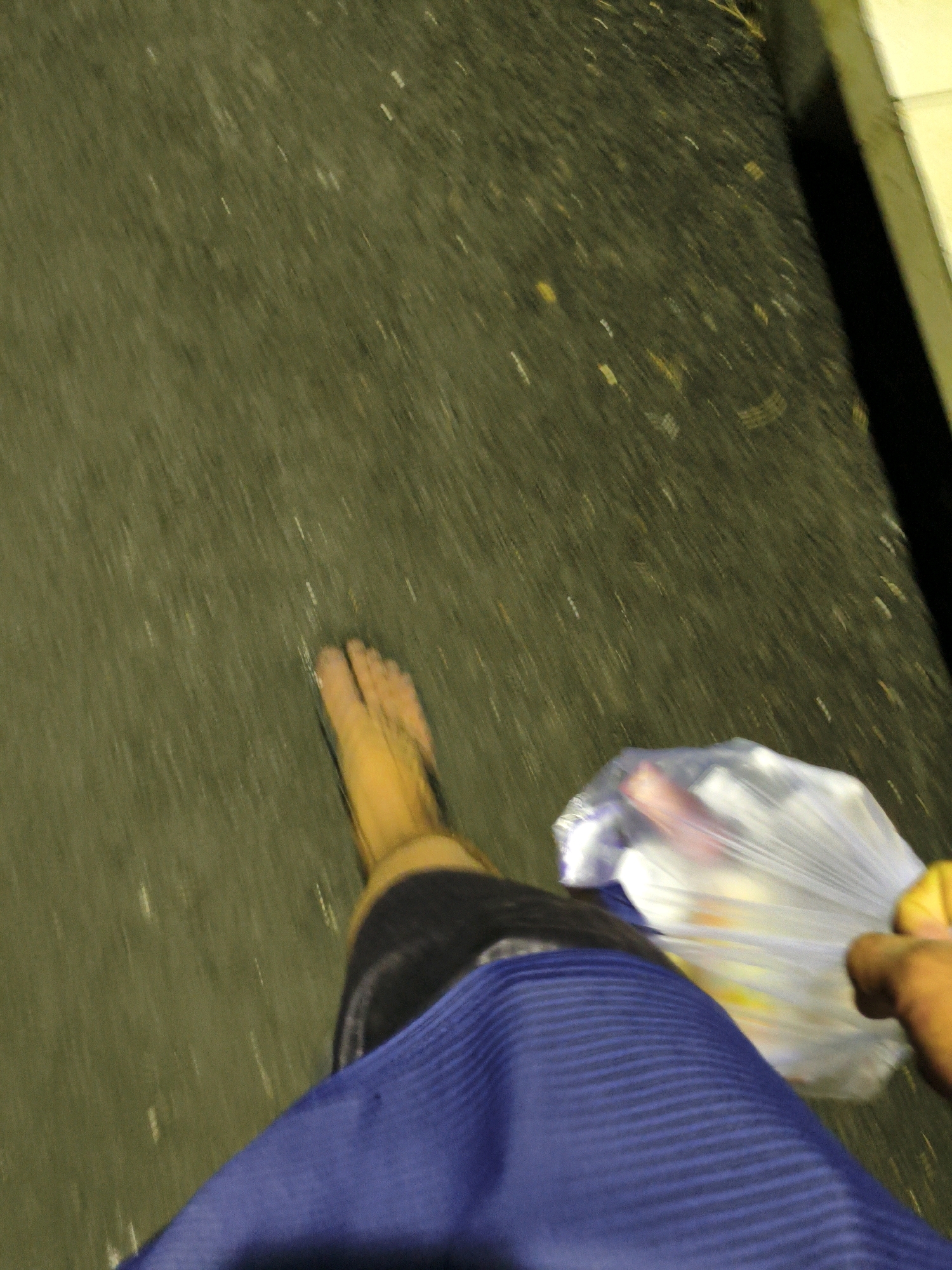 The image shows a person walking barefoot on a dark, textured surface, possibly asphalt, with some scattered debris. The person is wearing a blue garment that appears to be a skirt or sarong. In their right hand, they are holding a transparent plastic bag filled with objects, some of which are brightly colored. The lighting suggests it is either nighttime or a very dimly lit area. The composition and angle suggest the photo was taken from a moving perspective, possibly by the person themselves or someone walking alongside them. There are no identifiable landmarks to confirm the location in Bekasi, Indonesia, but the general urban environment implied by the asphalt and debris is consistent with such a location.