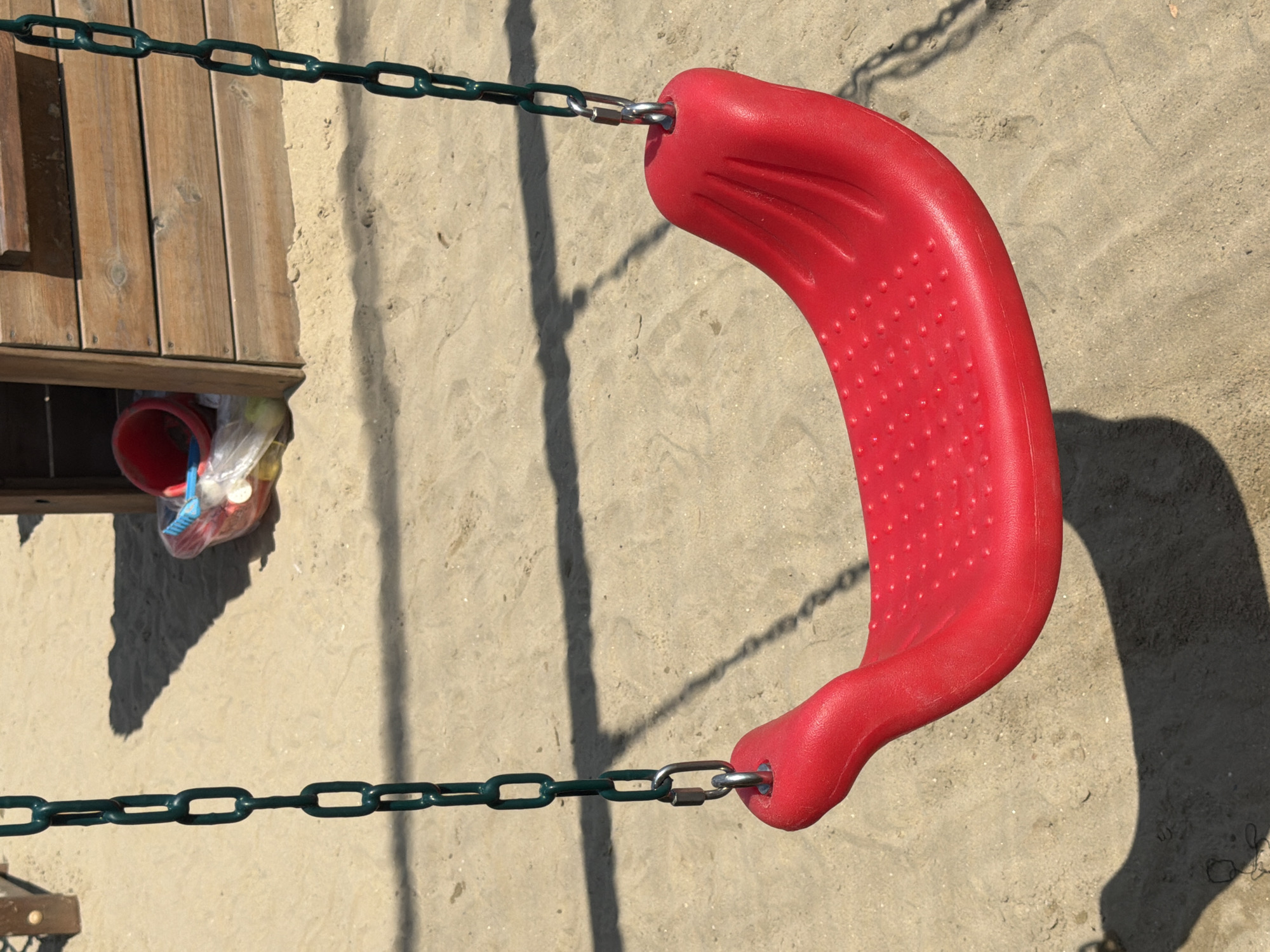 This image depicts a red swing seat hanging from dark green chains in a sandy playground. The swing is empty. In the background, part of a wooden play structure is visible, and a red bucket with some toys inside is placed on the sand near the structure. The ground is covered in light-colored sand with visible footprints and shadows cast by the swing and play structure, indicating it is daytime. The scene appears to be located outdoors, likely in a public park or playground. The location is identified as Gatteo, Italy.