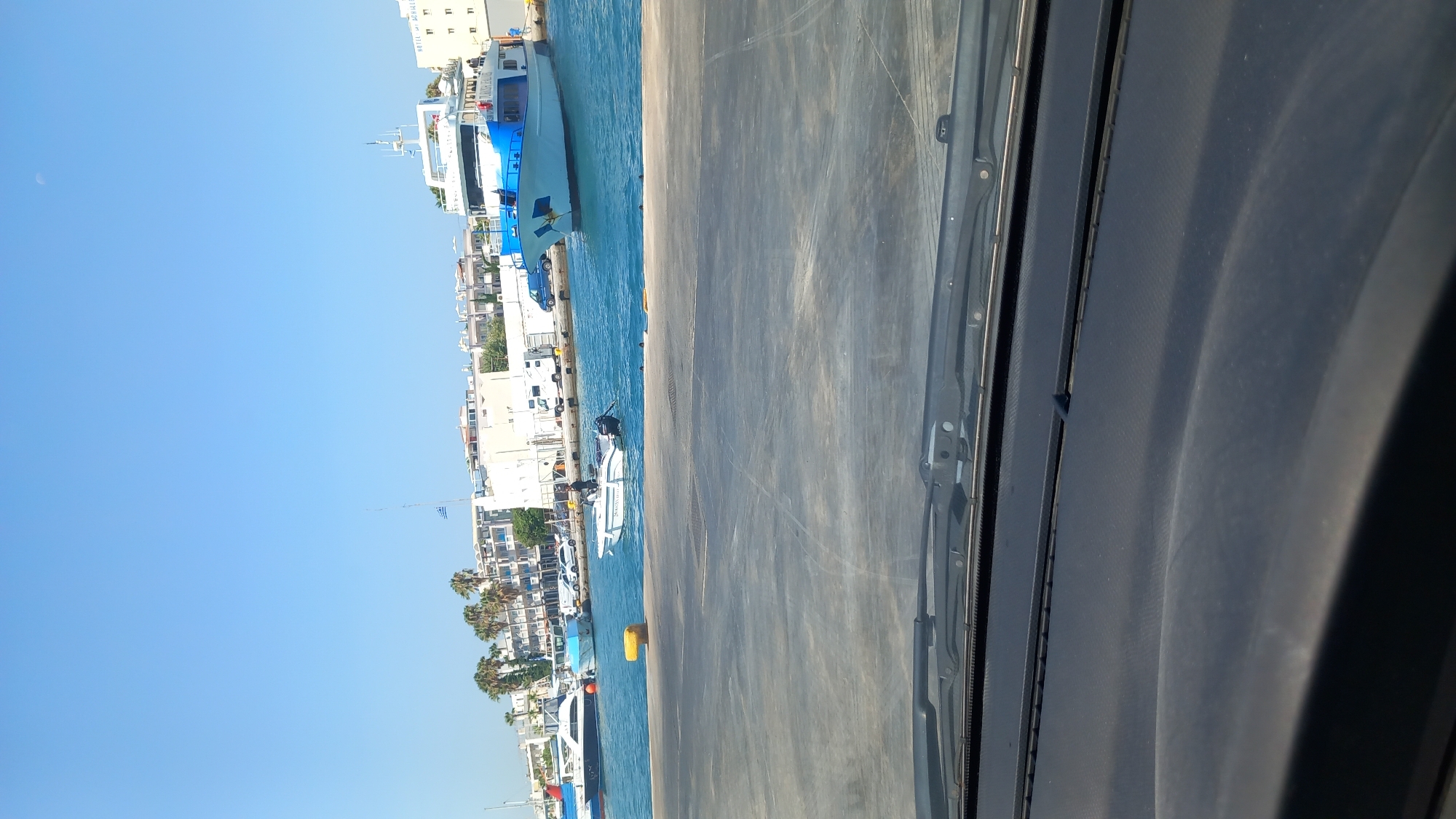 The image captures a view from a vehicle's dashboard, looking out over a harbor in the city of Kos, Greece. The foreground is dominated by the dark, textured surface of the car's dashboard and windshield wipers. Beyond the vehicle, the clear blue water of the harbor is visible, dotted with various boats. A prominent ferry, painted white with a blue upper deck and railing, is docked on the right side of the frame. The ferry has the name "TÜRKSUN" written on its side in blue lettering. Several other smaller boats, including a white speedboat with an outboard motor and a white yacht, are moored in the water. On the shore, buildings with light-colored facades, palm trees, and a Greek flag flying from a tall flagpole are visible. The sky is a vibrant, cloudless blue. A crescent moon is faintly visible in the upper right corner of the sky. The overall scene depicts a sunny day at a bustling harbor.