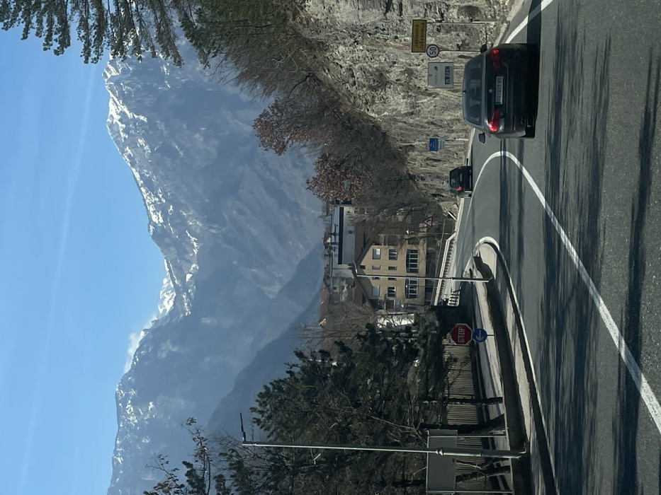 The image depicts a daytime scene in Jablanica, Bosnia and Herzegovina, specifically along a two-lane asphalt road curving slightly to the left. In the foreground, a dark-colored sedan, a BMW with a license plate partially visible as "T09-M-110", travels away from the viewer in the right lane. Further ahead in the left lane, a dark-colored hatchback or small SUV also moves away from the viewer.
To the right of the road, a steep, rocky cliff face with sparse, bare deciduous trees and evergreen shrubs ascends. Road signs are affixed to the cliff face: a yellow rectangular sign reading "Jablanica Јабланица" above a circular 50 km/h speed limit sign, followed by a yellow "PLAC" sign with a right-pointing arrow. Below these are blue rectangular highway signs indicating "E73" and "M17", and another blue sign with an arrow pointing to "Mostar".
To the left of the road, a metal guardrail or fence runs parallel to the asphalt, behind which are several evergreen trees and bare deciduous trees. A street light pole is visible on the left. Further down the road, past the trees, a red octagonal "STOP" sign is posted, accompanied by a blue circular sign featuring a white left-pointing arrow. Behind these signs and trees, multi-story buildings belonging to the town of Jablanica are visible, including a prominent light-colored building with multiple windows and a reddish-brown roof.
In the background, under a clear blue sky, majestic mountains with visible snow patches on their peaks dominate the horizon, creating a mountainous valley setting for the town. The overall lighting suggests bright sunlight, casting shadows on the road and structures.