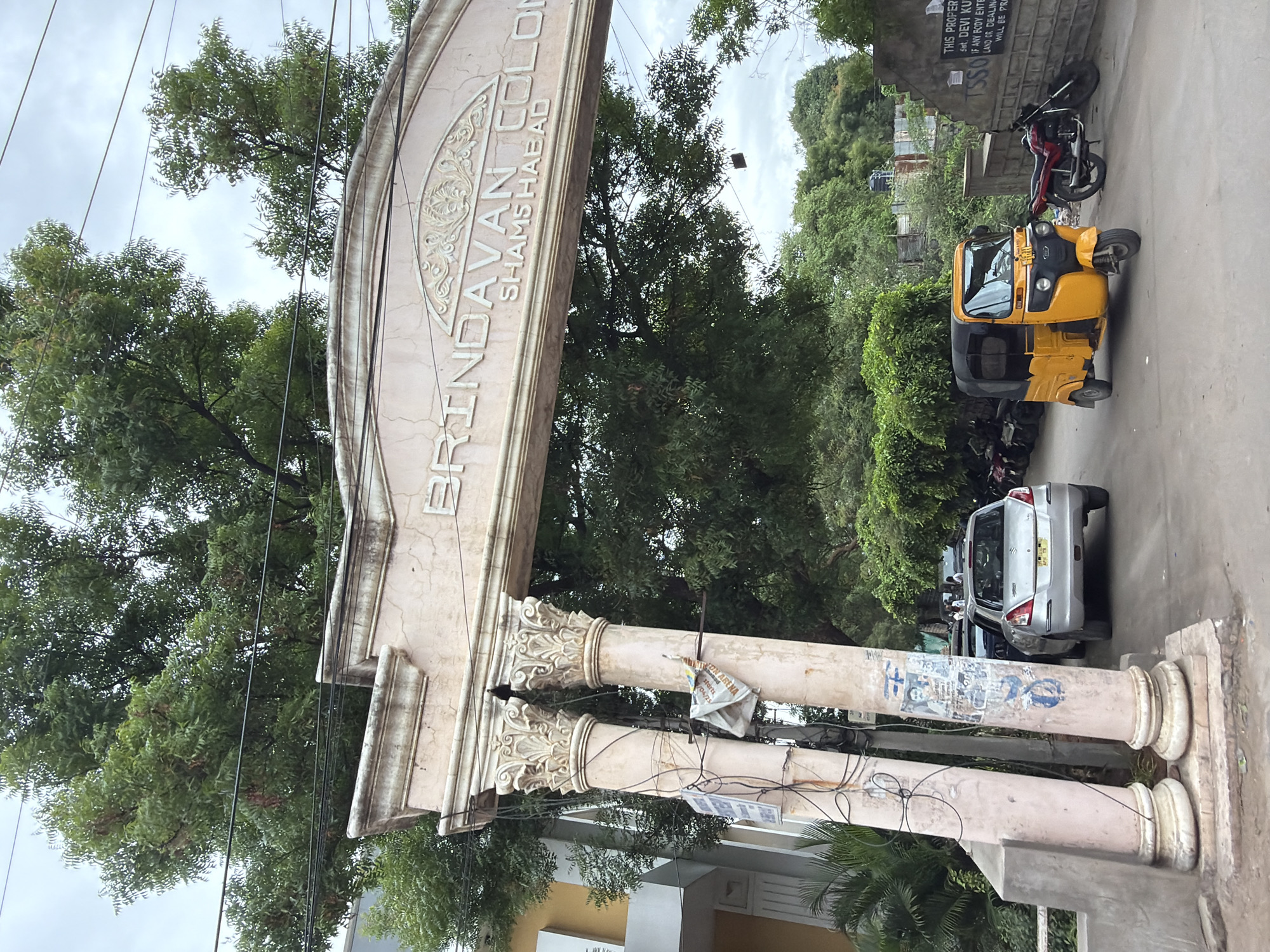 The image captures the entrance archway to "BRINDAVAN COLONY SHAMS HABAD" in Shamshabad, India. The archway, made of stone, is supported by two ornate pillars. Wires and posters are attached to the pillars. Beyond the archway, a silver hatchback car is parked on the left, and a yellow auto-rickshaw is visible in the center. A red motorcycle is parked to the right of the auto-rickshaw, leaning against a stone wall. Several other motorcycles are parked further down the road on the left, behind the car. Lush green trees form a canopy over the scene, with a cloudy sky in the background. A sign is affixed to the stone wall on the right. The overall scene depicts a residential colony entrance with vehicles and foliage typical of an urban Indian setting.