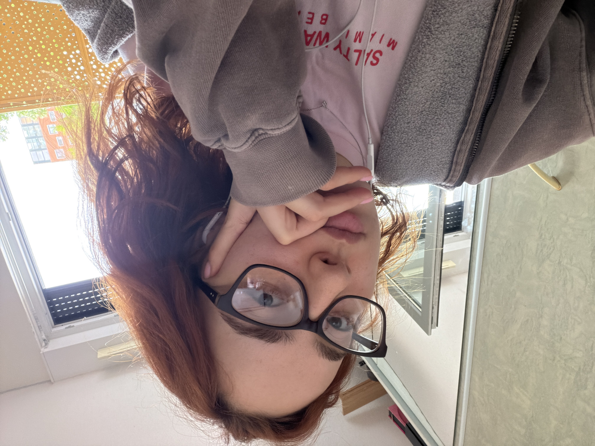 A close-up selfie shows a young person, likely female, with reddish-brown hair, looking directly at the camera through black-framed glasses. The image is taken from a low angle, looking upwards towards the subject. One hand is partially covering the left side of her face, with fingers obscuring her eye and pink-painted fingernails visible. Her lips are slightly parted, revealing a hint of her tongue.
She is wearing a light pink t-shirt with red text that reads "SALTY WA M" and "MIAMI BE" (partially cut off, suggesting "Salty Waves Miami Beach"). Over the t-shirt, she wears a grey zippered hoodie. A white earbud wire hangs from her ear, disappearing under her clothing.
The background is brightly lit, primarily by a large window. Outside the window, a multi-story building with reddish-orange brick walls and several windows is visible. Above the window, an exterior perforated gold-colored screen or awning with circular cutouts casts speckled light. On the right side of the frame, a reflective surface, possibly a mirror or a cabinet, is visible, reflecting some light and indistinct shapes. The bright light suggests it is daytime. The overall setting appears to be indoors, near a window in a room in Strasbourg, France.