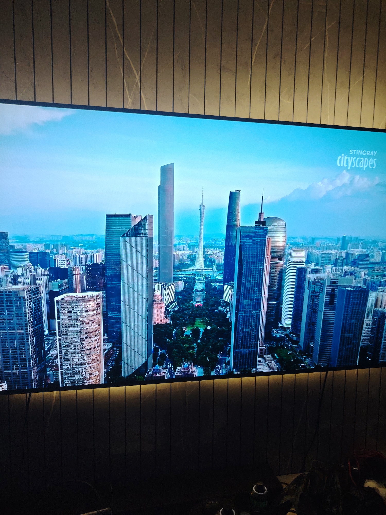 The image captures a widescreen display showcasing a panoramic view of a modern cityscape, set within an indoor environment. The screen presents an aerial perspective of numerous skyscrapers and high-rise buildings under a clear blue sky with some scattered white clouds. Key architectural features on the screen include a very tall, slender dark skyscraper on the right, and a prominent, distinctively shaped tower with a lattice structure, identifiable as the Guangzhou Tower, on the far left. Green park areas are visible interspersed among the buildings. In the upper right corner of the screen, the white text "STINGRAY" appears above "cityscapes."
The screen is bordered by interior architecture. Above the display, a wall segment features vertical panels with a light brown, marble-like pattern, exhibiting subtle darker veining. Below the screen, a dark, horizontal surface is visible, possibly a counter or furniture, with faint reflections of light. In the lower left corner, two white cylindrical objects, possibly cups, are partially visible, and in the lower right, an indistinct dark object with some reddish accents and fabric texture is present. The overall setting, including the display and its immediate surroundings, is located in Huntingdonshire, United Kingdom.