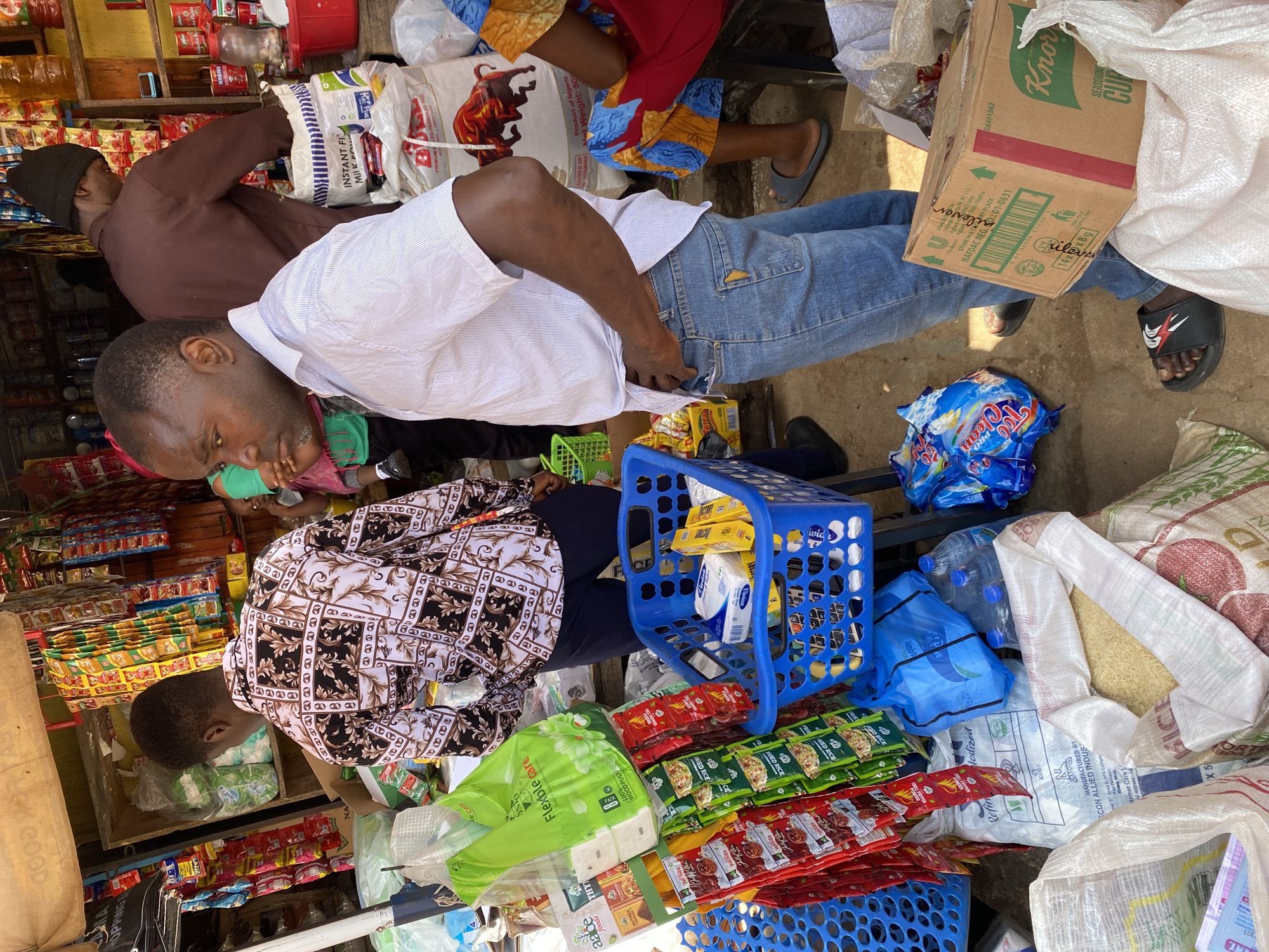 This is a medium shot of a bustling market stall in Jos, Nigeria. The foreground features a man in a blue striped shirt and denim jeans, with his hands on his hips. To his left, a blue plastic basket overflows with goods, including what appear to be packs of tissues and yellow boxes. Beside the basket, several bags of rice and other dry goods are piled on the ground, along with a blue plastic bag and several bottles of water. To the left of the man, a display rack is filled with small packets of various food items. In the background, another man in a brown long-sleeved shirt and a black beanie is attending to goods on a shelf. The stall is packed with merchandise, including bags of flour or rice, bottles of oil, and numerous packaged snacks and food items hanging from the ceiling and shelves. The overall impression is one of a lively and well-stocked market environment.