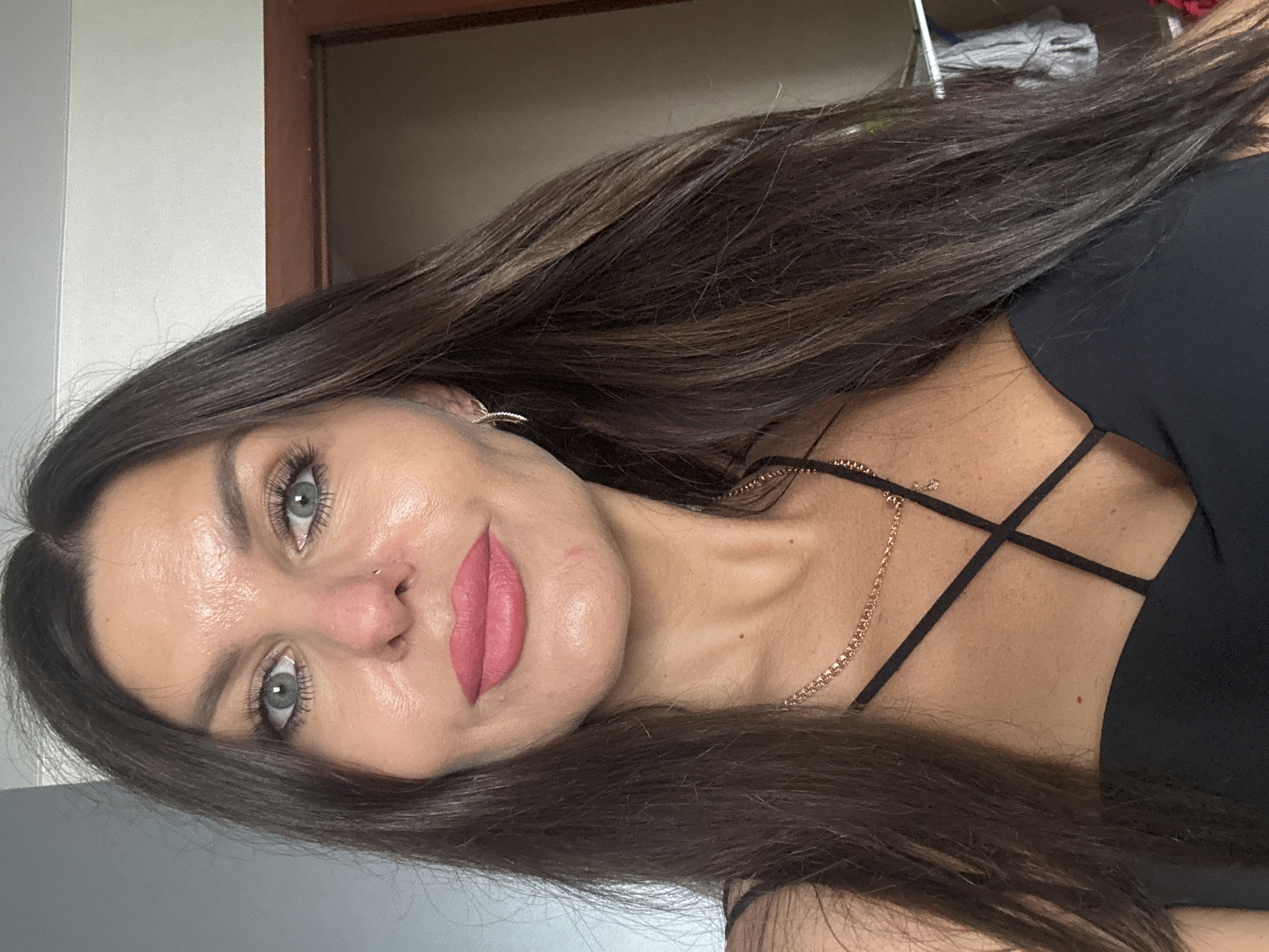 A medium shot portrait of a woman with long, dark brown hair framing her face. She has striking blue-green eyes, defined eyebrows, and a small nose stud. Her lips are painted a matte pink. She is wearing a black top with thin, crisscrossing straps across her chest and a rose gold chain necklace with a small pendant. A subtle glint of a hoop earring is visible on her left ear. The background shows a wall and a doorway, suggesting an indoor setting, possibly a home. There is no information about the city of Chaikovsky, Russia, present in the image.