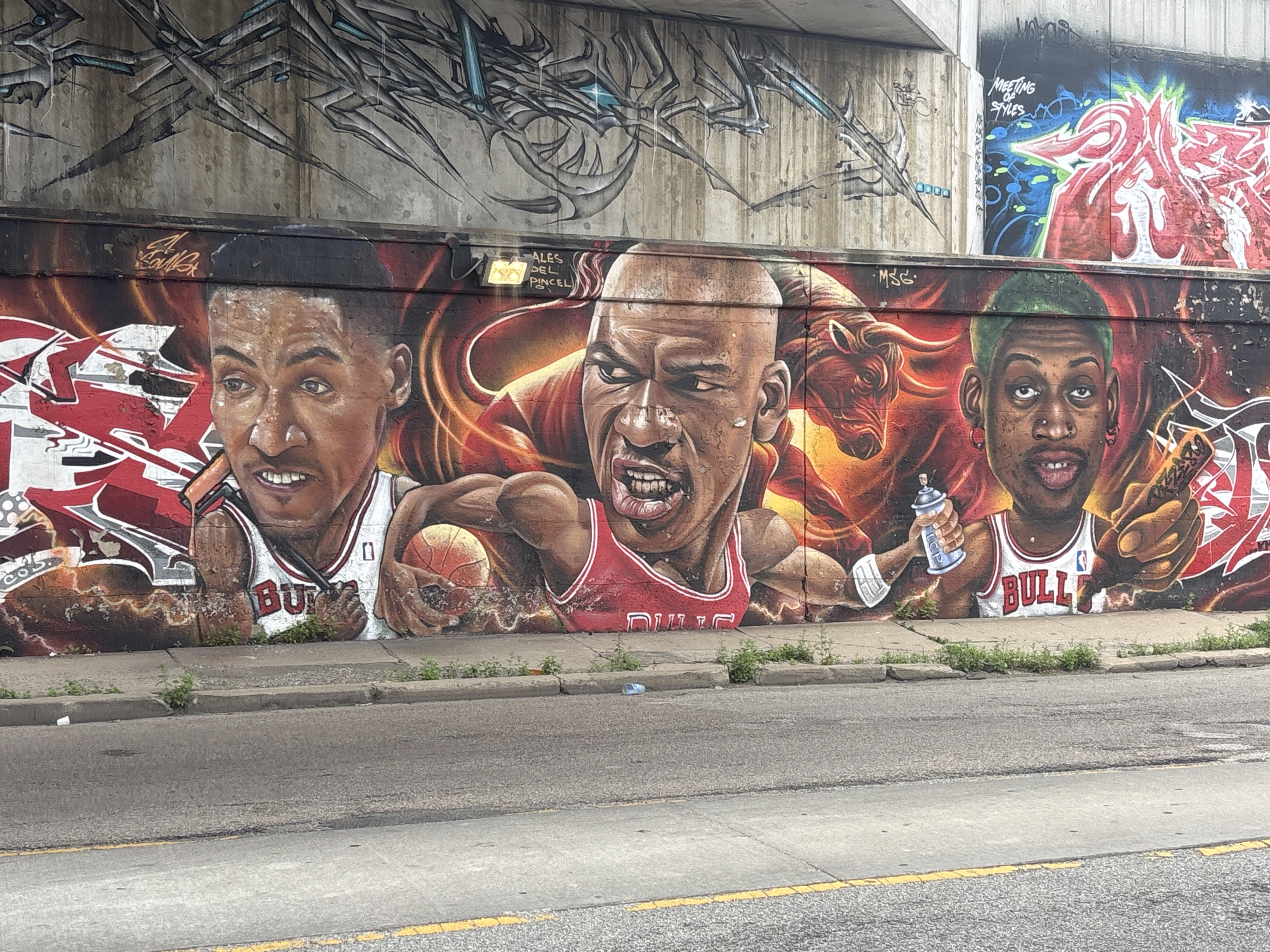 The image shows a mural on a concrete wall in Chicago, United States of America. The mural depicts three Chicago Bulls players: Scottie Pippen, Michael Jordan, and Dennis Rodman. Scottie Pippen is on the left, Michael Jordan is in the center, and Dennis Rodman is on the right. Michael Jordan is holding a basketball and appears to be dribbling it. Dennis Rodman is holding a spray can. Behind them is a fiery red and orange background with a bull's head on the left side. Above the mural, there is more graffiti on the concrete wall. The mural is located outdoors, adjacent to a street with a sidewalk. There is some debris on the sidewalk and street.