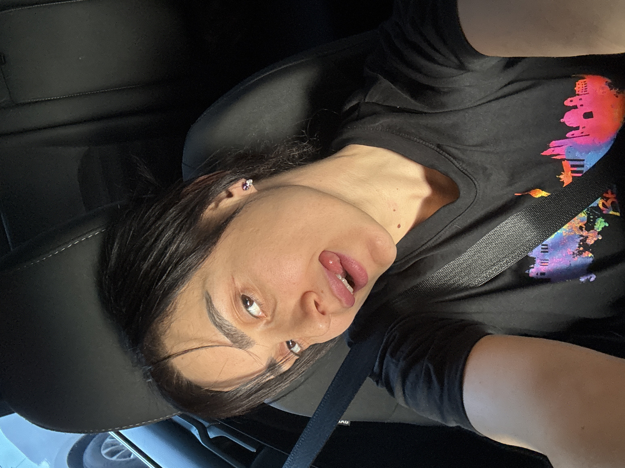 A woman is seated in a car wearing a black t-shirt with a colorful graphic print. She is wearing a seatbelt and has dark hair and visible earrings. Her tongue is sticking out slightly. The location appears to be the city of Tbilisi, Georgia as the t-shirt has the skyline of Tbilisi on it. The interior of the car is mostly black, and a glimpse of a tire is visible in the lower left corner.