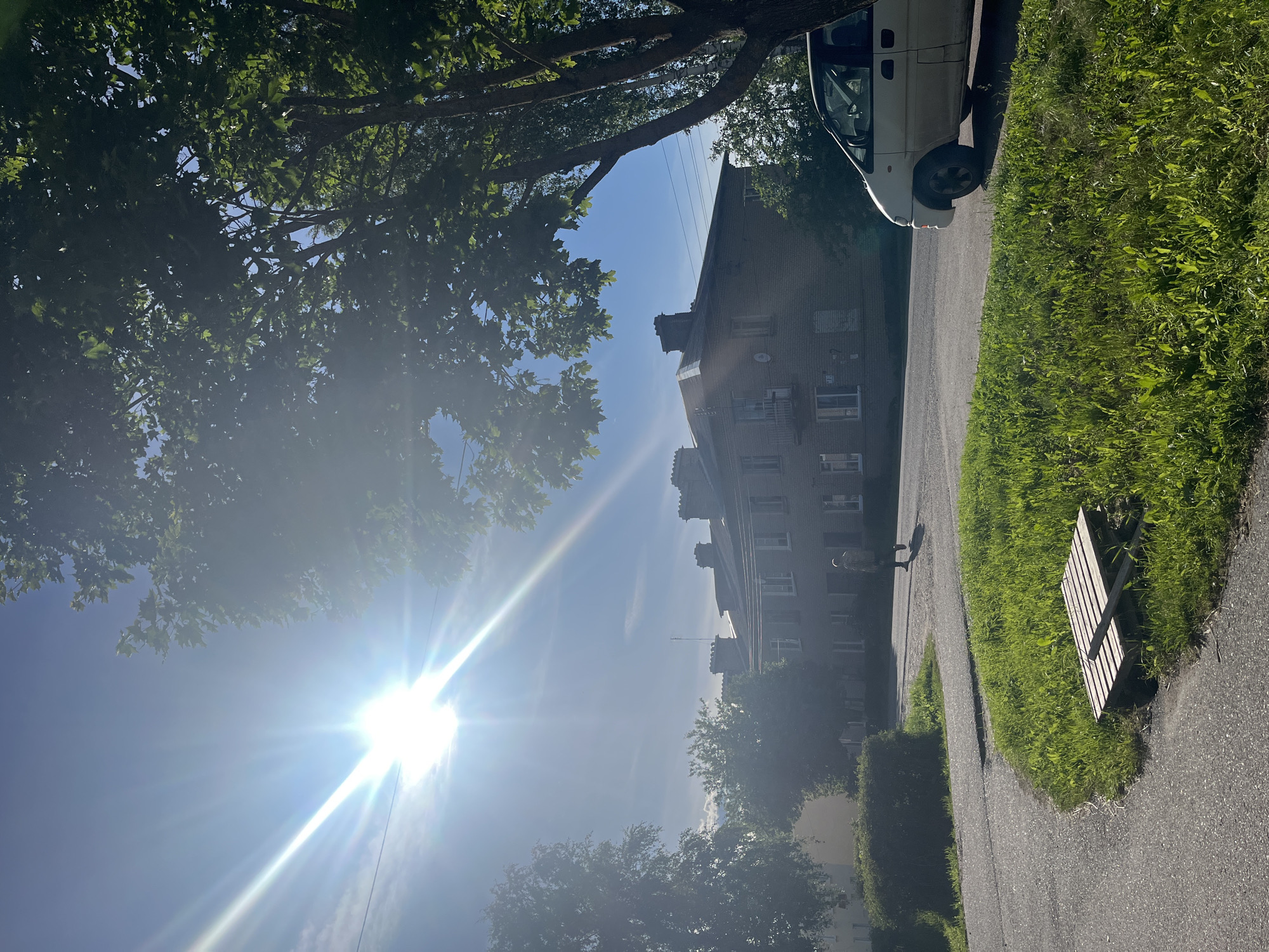 The image depicts an outdoor scene in a residential area, likely in Серебрянский, Russia. A bright sun, positioned low in the sky, casts lens flare across the frame, indicating it's either early morning or late afternoon. The sky is a clear blue, suggesting fair weather.
The foreground features a paved pathway bordered by lush green grass and a wooden bench. To the left, the branches of a large deciduous tree with vibrant green leaves create a canopy, partially obscuring the sky. Beyond the tree, a multi-story brick apartment building is visible. The building has several windows and chimneys, hinting at older architecture.
A white van is parked on the left side of the pathway, close to the apartment building. A solitary figure, appearing to be a person, is walking on the pathway in the middle distance, casting a shadow due to the strong sunlight. The overall atmosphere is serene and peaceful, conveying a sense of a typical day in a neighborhood. The presence of trees, a residential building, and a pathway suggest an urban or suburban setting. No visible text is present in the image.