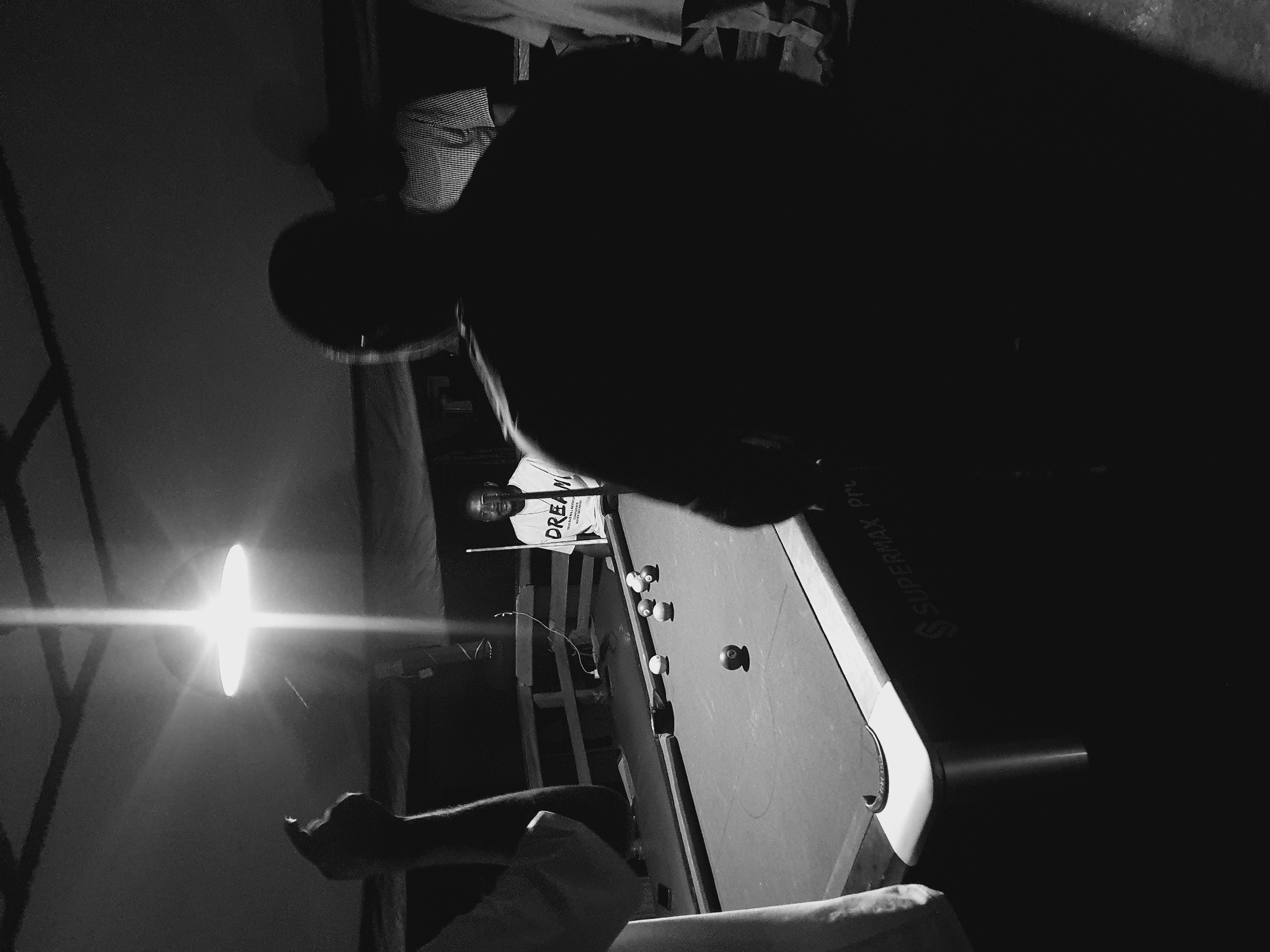This is a black and white, low-light photograph of a group of people playing pool. The scene appears to be outdoors, possibly in a makeshift establishment in Lokoja, Nigeria, indicated by the presence of a "Supermax Pro" pool table, a brand common in such settings. A bright overhead light illuminates the pool table and some of the individuals.
In the foreground, the back of a person is visible, silhouetted against the light, obscuring their face. To the left, another person's arm is raised, with a finger pointing upwards, possibly indicating a shot or making a gesture.
In the mid-ground, a man wearing a white t-shirt with the word "DREAM" printed on it is standing behind the pool table, holding a cue stick. He is looking towards the viewer with a slight smile. Several pool balls are scattered on the table, some in play and others in position.
To the right of the man in the "DREAM" shirt, another person is partially visible, wearing a striped shirt. Further to the right, the legs of another person are visible, suggesting more people are present in the scene, either observing or participating in the game.
The environment seems rustic, with a wooden railing or fence visible behind the pool table. The overall atmosphere is casual and social, depicting a moment of recreation among friends. The low light and high contrast suggest the photograph was taken at night or in a dimly lit area.